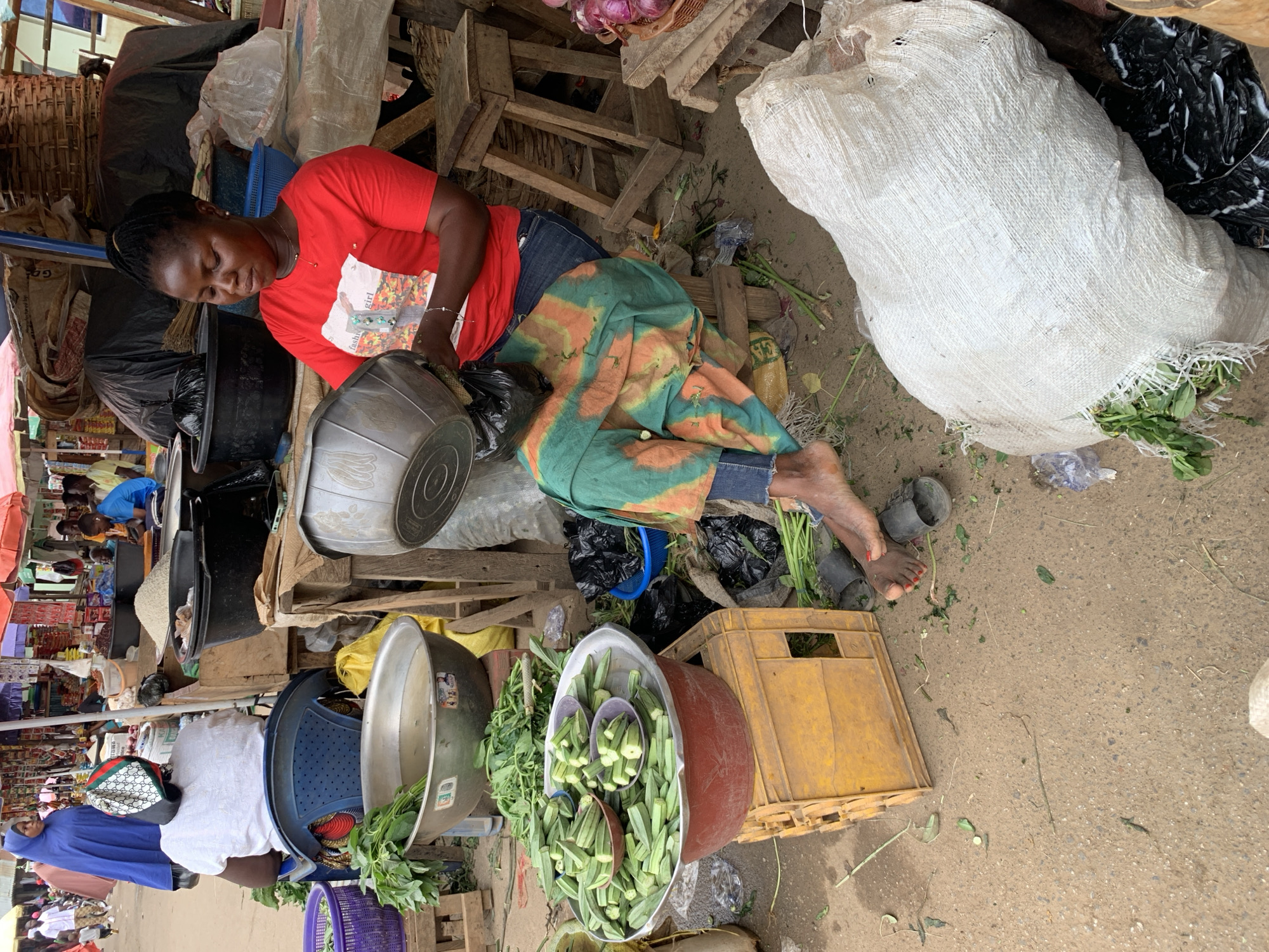 This image captures a bustling outdoor market scene in Akwanga, Nigeria, characterized by makeshift stalls and a vibrant display of fresh produce. The ground is unpaved, appearing as dirt or sand.
In the foreground, a woman with dark skin and braided hair is the central figure. She wears a red t-shirt with a graphic (partially visible, possibly "fashion girl"), blue jeans, and a patterned green and orange wrapper or skirt over her legs. She is sitting on a low wooden structure or stool, intently focused on a task involving a metal basin and black plastic bags, likely sorting or preparing goods for sale. Her bare feet, one with a sandal nearby, are visible on the ground.
Surrounding her and extending into the background are numerous items typically found in a market. Large quantities of fresh produce are prominent, including a large red basin filled with green okra pods, with smaller bowls of okra nestled within it. Nearby, green leafy vegetables are stored in metal bowls and plastic baskets. A large white sack, possibly containing more produce or commodities, with some greens spilling from it, rests on the ground. Onions can be seen in a basket in the upper right.
Various containers such as metal basins, plastic crates (one yellow), blue and purple plastic baskets, and black plastic bags are scattered throughout the scene. Wooden planks and makeshift structures form parts of the vendor stalls.
In the background, other market-goers and vendors are visible. One person in a blue robe stands to the left, while another person in a white shirt and a distinct Gucci-branded cap is seen from behind. The natural lighting suggests it is daytime. The overall atmosphere indicates active commerce and daily life in the market.