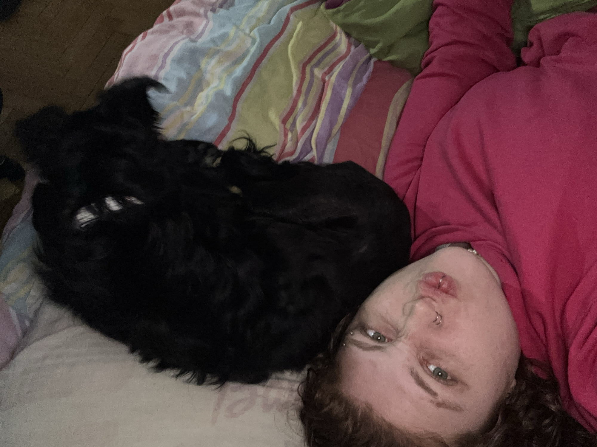 A woman with curly red hair and facial piercings, wearing a red sweatshirt, lies on a bed with striped bedding. A black dog is curled up beside her. The location is unspecified; there is no information to suggest this image was taken in Buenos Aires, Argentina.