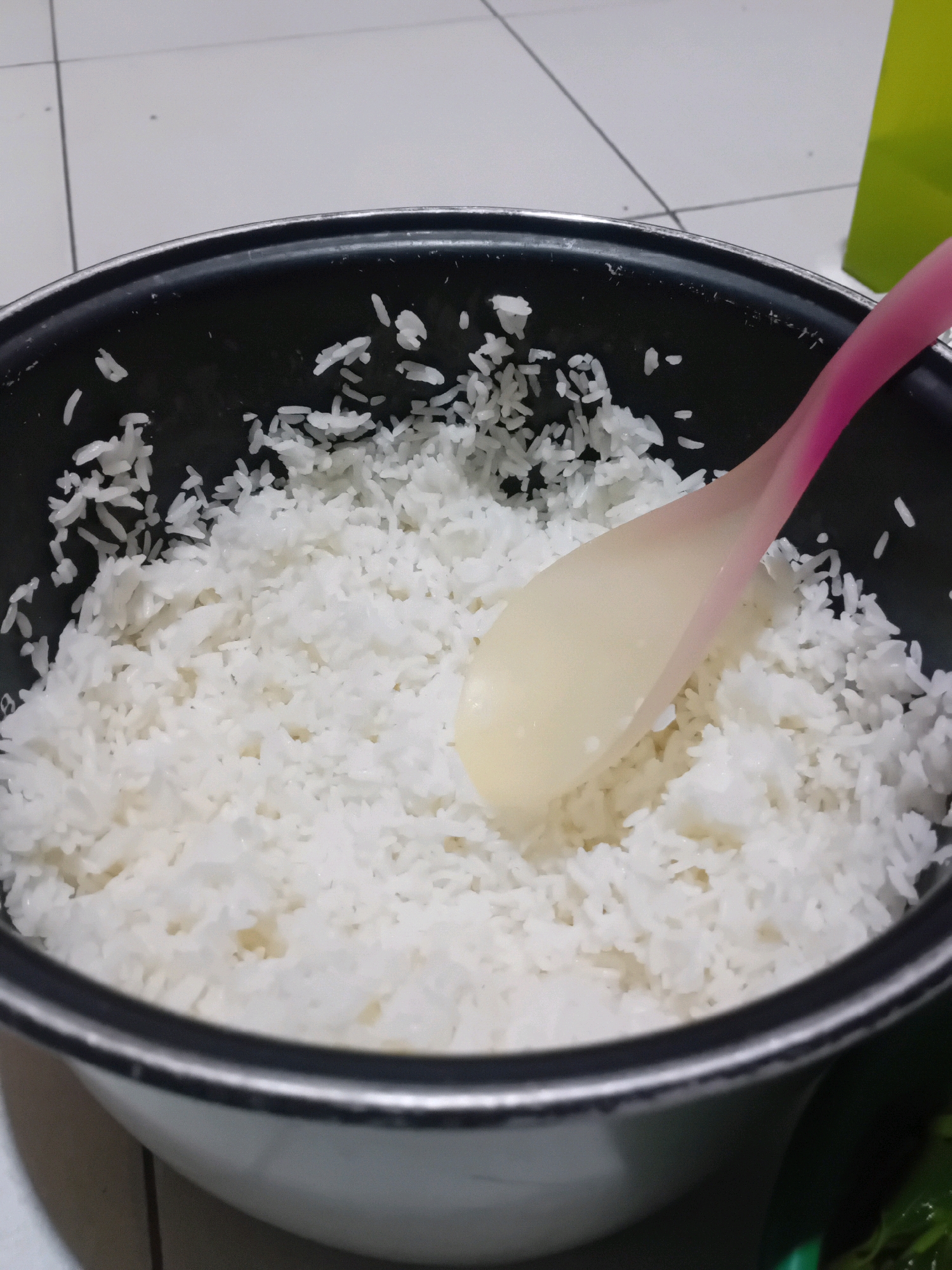 A close-up, high-angle shot shows a rice cooker filled with freshly cooked white rice. A translucent pink and yellow rice paddle rests on top of the rice, partially submerged. The rice cooker is black with a silver rim. The background is out of focus, but appears to be a tiled floor and a bright green object, possibly a bag. The scene is likely domestic, and the image was taken in Pekanbaru, Indonesia.