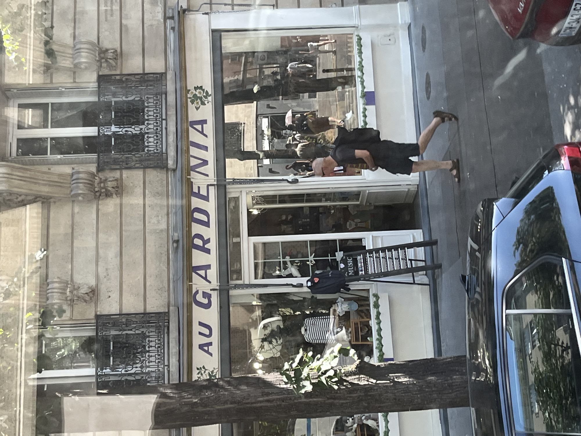 The image depicts the facade of a shop in Paris, France, identified by the signage "AU GARDENIA." The shop has a large display window showcasing various items, including clothing on mannequins and a display stand labeled "MUSIC." Outside the shop, a man with a backpack walks past, heading away from the camera. The street in front of the shop is visible, along with reflections in the shop window showing other pedestrians and buildings across the street. The architecture suggests a classic Parisian style, with ornate balconies on the upper floors of the building. A black car with a shark fin antenna is parked in the foreground on the left, and part of a red car is visible on the right. The time of day appears to be daytime, with sunlight casting shadows.