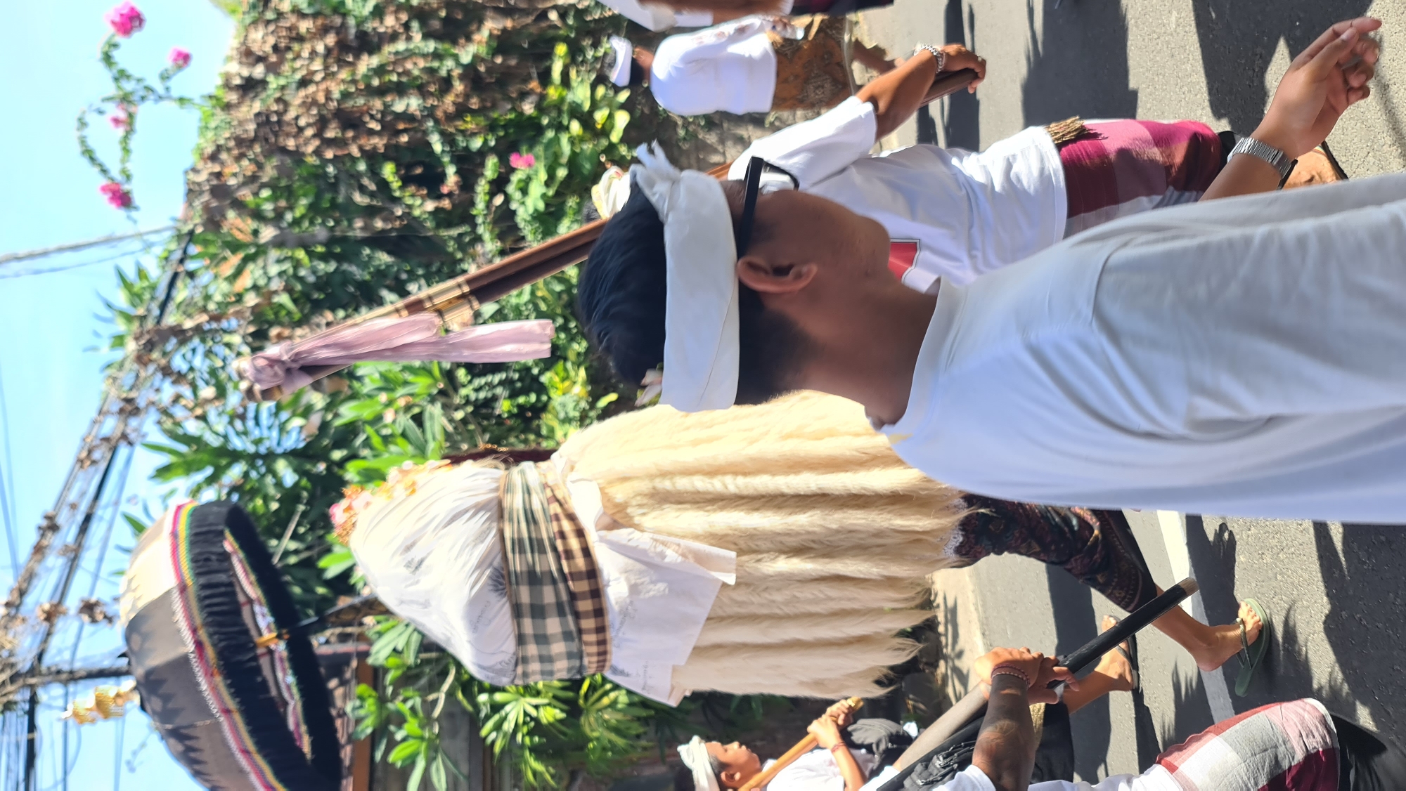 This image captures a street scene in Denpasar, Indonesia, likely during a cultural or religious procession. In the foreground, a person wearing a white t-shirt and a white headband with glasses is seen in profile, walking to the right. Behind them, a figure adorned with a tall, conical headdress covered in white cloth and a checkered sash is partially visible. This figure is also draped in a cream-colored, shaggy material. To the left, another person in a white t-shirt is playing a wooden flute-like instrument. Further in the background, more individuals in white shirts and patterned lower garments, typical of Balinese attire, are walking. An ornate umbrella, likely part of the procession, is held aloft, decorated with colorful trim and fringe. The environment features lush green foliage and a vine with pink flowers climbing over a wall or structure, suggesting an outdoor, possibly tropical setting. Overhead, power lines crisscross the bright blue sky. The street surface appears to be asphalt. The overall impression is one of a lively cultural event taking place outdoors.