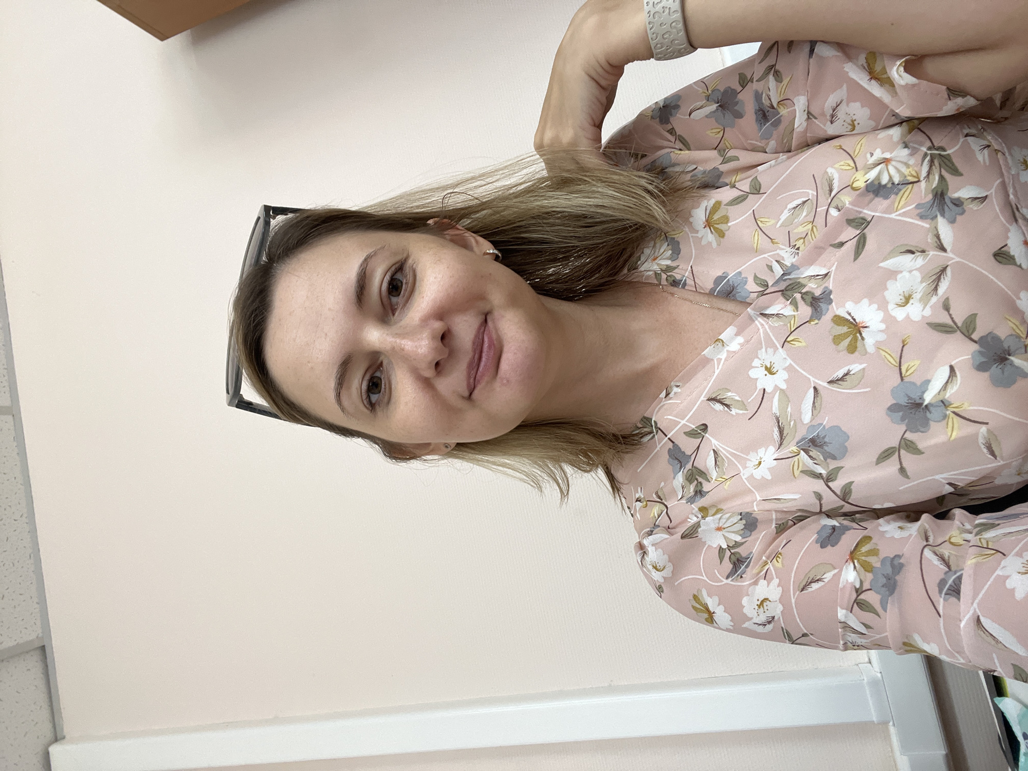A medium-shot selfie shows a woman in her 30s from the chest up, posing indoors against a pale pink wall. She has fair skin, light brown hair that falls to her shoulders, and brown eyes. She is wearing a light pink V-neck top with a floral pattern of white, gray, and yellow flowers and green leaves. Her gaze is directed at the camera, and she has a slight smile. She is wearing small hoop earrings and a watch on her left wrist. A pair of glasses is perched on top of her head. The background is plain, with a white vertical trim on the left and a hint of a brown object on the right. The scene appears to be an office or a domestic setting. There is no visible indication of the location being in Volgograd, Russia.