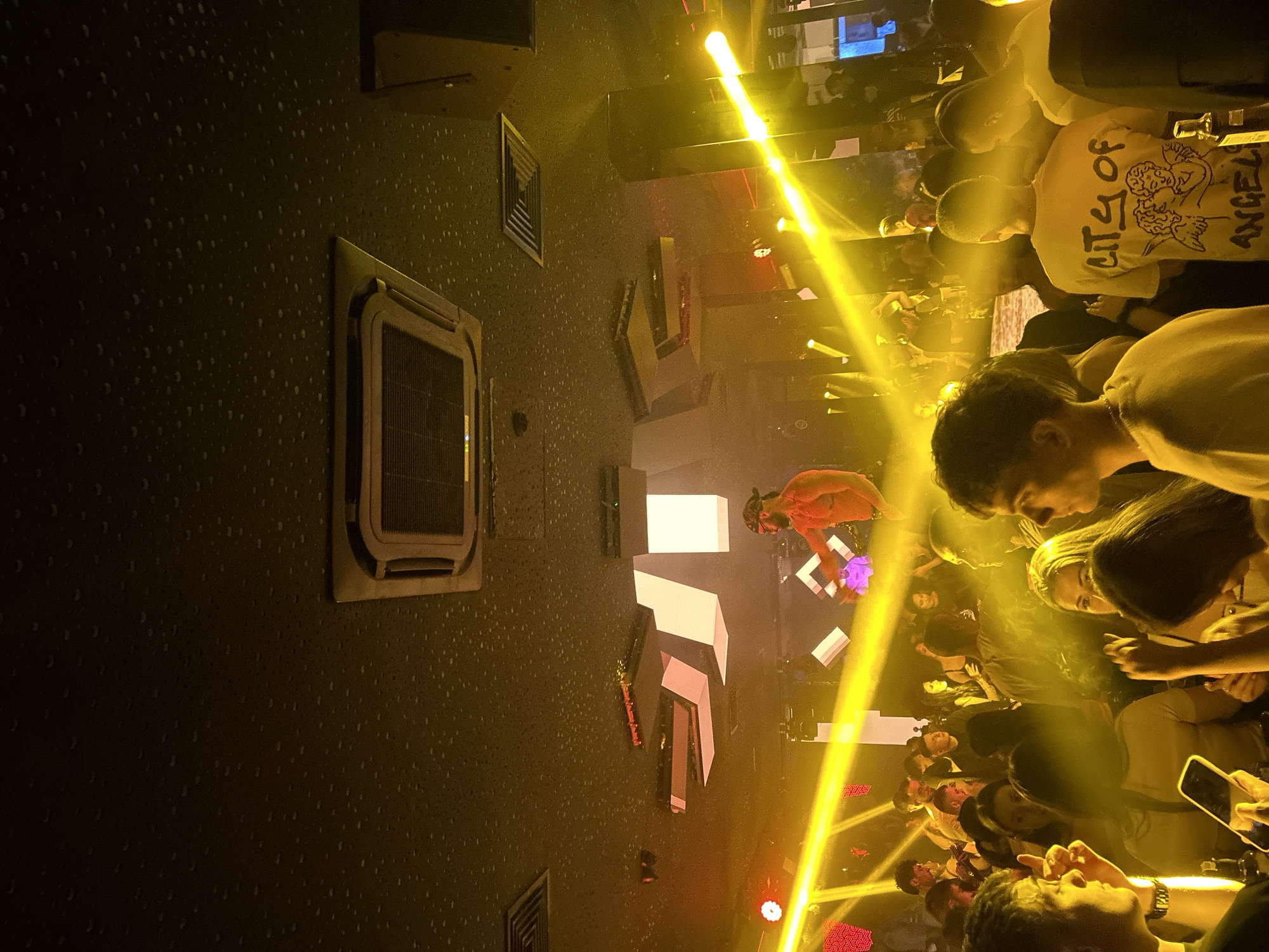 A nightlife scene is captured indoors, showing a DJ with a bare torso performing amidst a crowd. The location is a nightclub, possibly in Sofia, Bulgaria, suggested by the Cyrillic text visible in the background. The focus is on the crowded space with people in close proximity, many holding up phones, presumably recording the performance. Intense yellow lighting dominates the scene, with spotlights and illuminated panels adding to the vibrant atmosphere. There is visible ventilation and equipment on the walls. The foreground shows individuals partially visible, with attention directed towards the stage and the DJ.