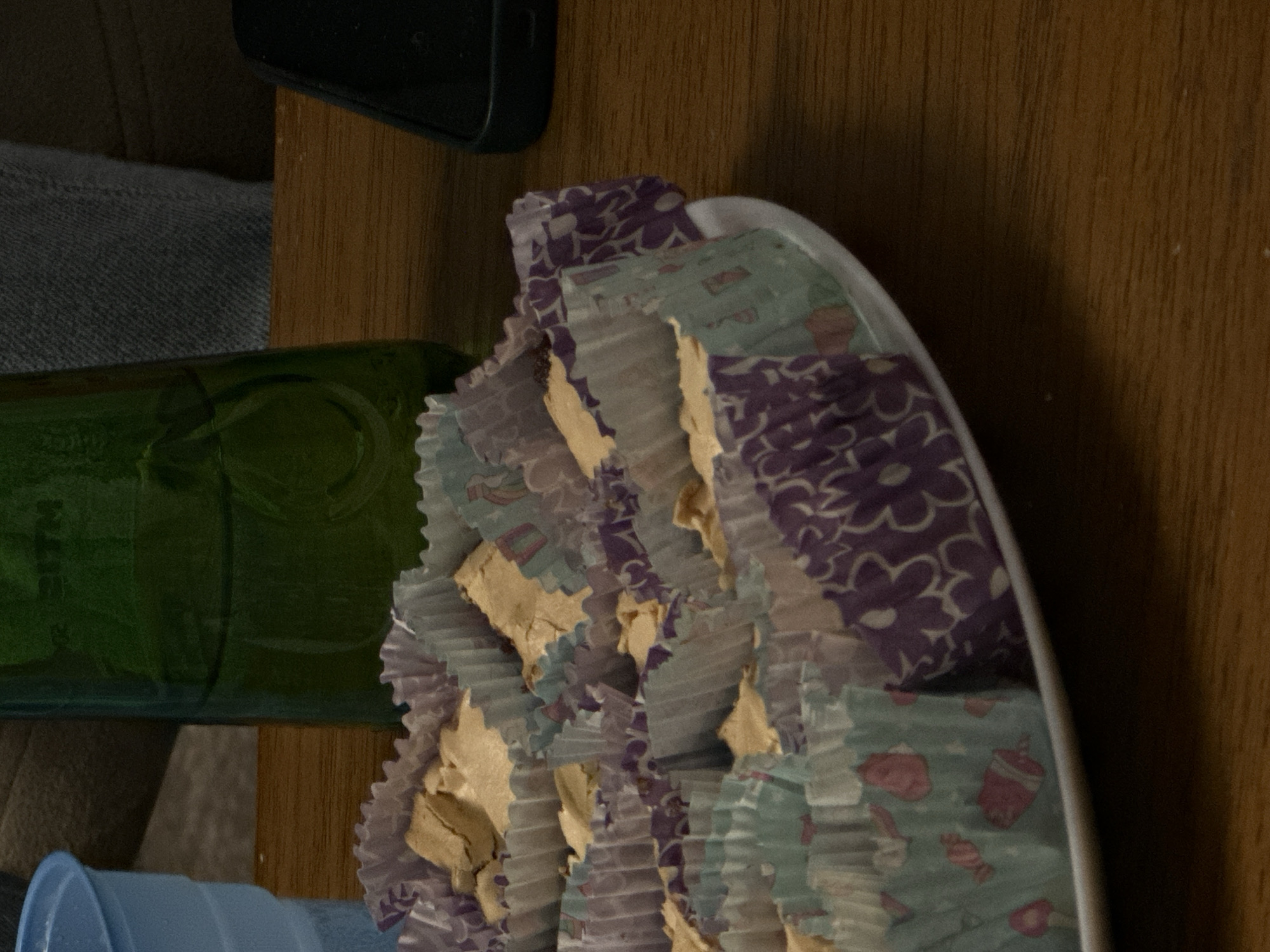 The image shows a partial view of a table setting indoors. Present are several items: A black smartphone is visible in the upper left corner of the frame. A green bottle lies horizontally, partially obscured by other objects. A stack of cupcakes in decorative paper wrappers is arranged on a white plate. A blue plastic container is situated in the lower left. The surface on which these objects are placed is a wooden tabletop with a visible grain pattern. The scene is lit with ambient light, though the exact source cannot be determined. There is no indication of location in the city of Tbilisi, Georgia. No people are visible, and no actions are taking place.