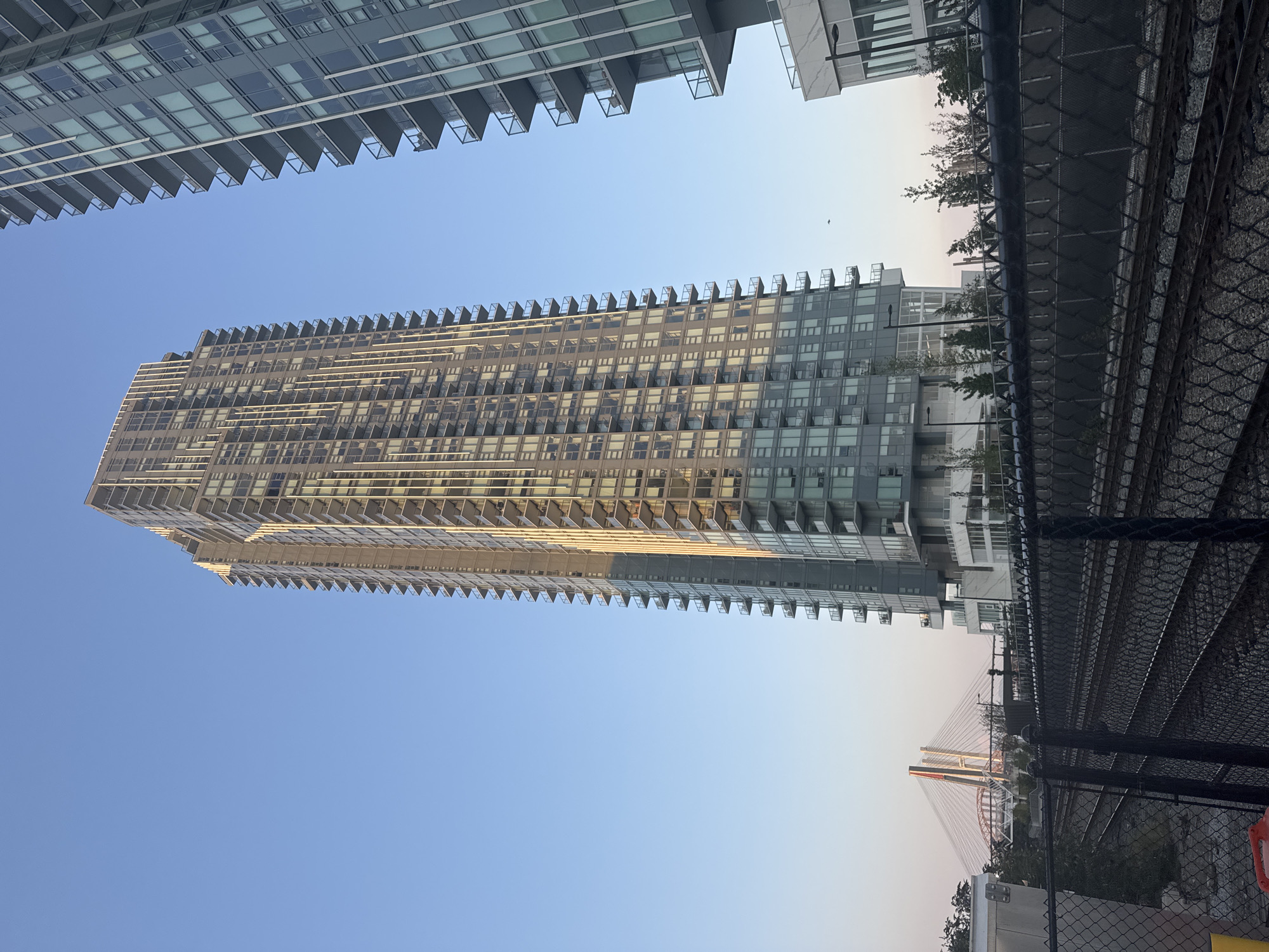 The photograph captures a view of a modern, high-rise residential building in New Westminster, Canada. The building, characterized by its numerous balconies and glass-paneled facade, dominates the frame under a clear blue sky. Adjacent to it, another similar building is partially visible on the right. In the foreground, railway tracks run horizontally across the image, separated from the viewer by a chain-link fence. Beyond the fence, some greenery and smaller structures are present. In the background, a suspension bridge with a distinctive red and white color scheme spans across a body of water, indicating the presence of the Fraser River. The lighting suggests it is either early morning or late afternoon, with the sun casting a warm glow on the side of the main building. There are no people visible in the image.