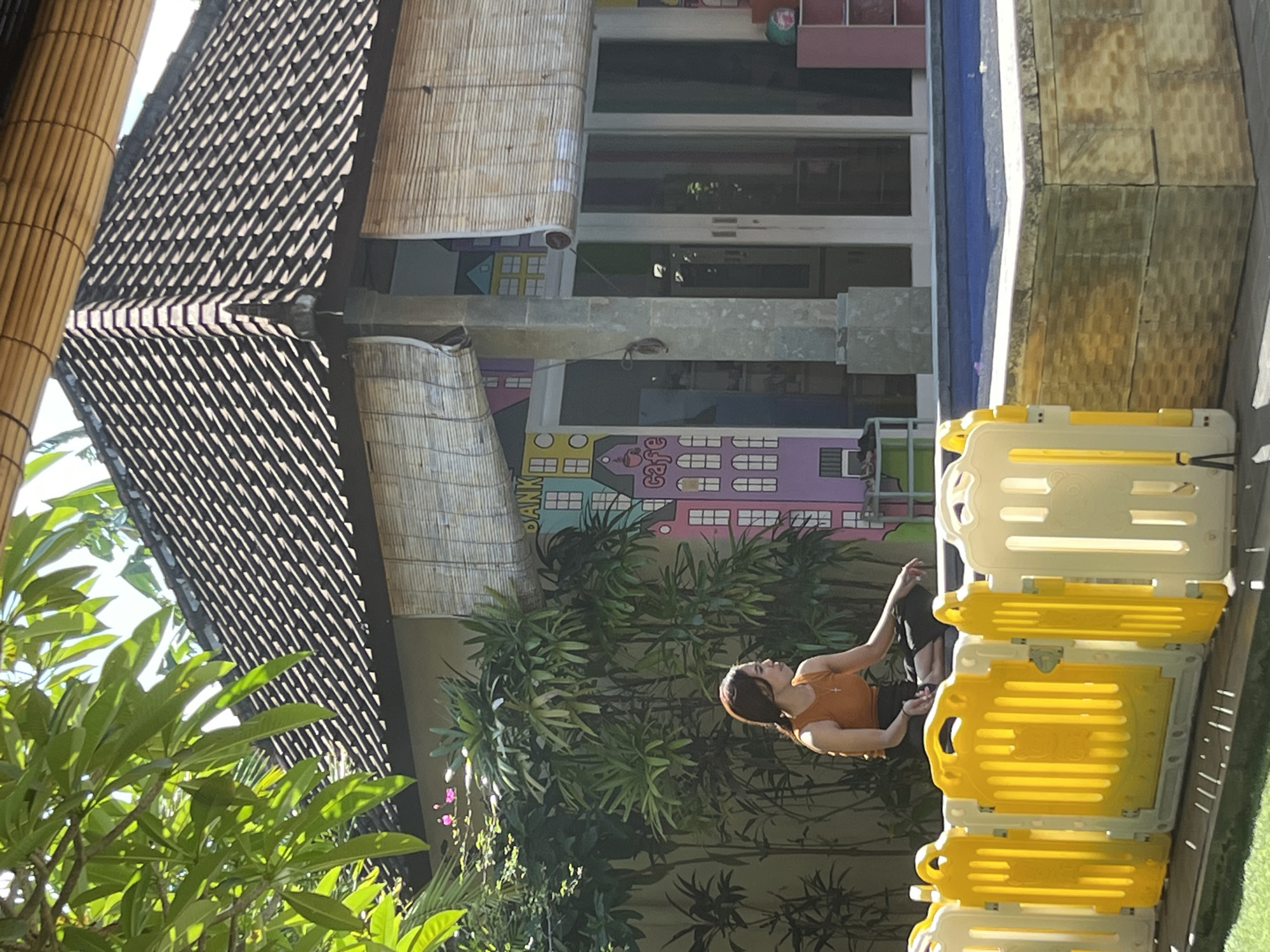 In Kuta, Indonesia, a young woman sits on the edge of a pool, facing left. She is wearing an orange tank top and black pants. The pool is surrounded by a wooden deck and a yellow and white plastic barrier. Behind the pool is a building with a tiled roof and bamboo blinds. The building is decorated with colorful murals of houses. Lush green foliage surrounds the building and the pool area.
