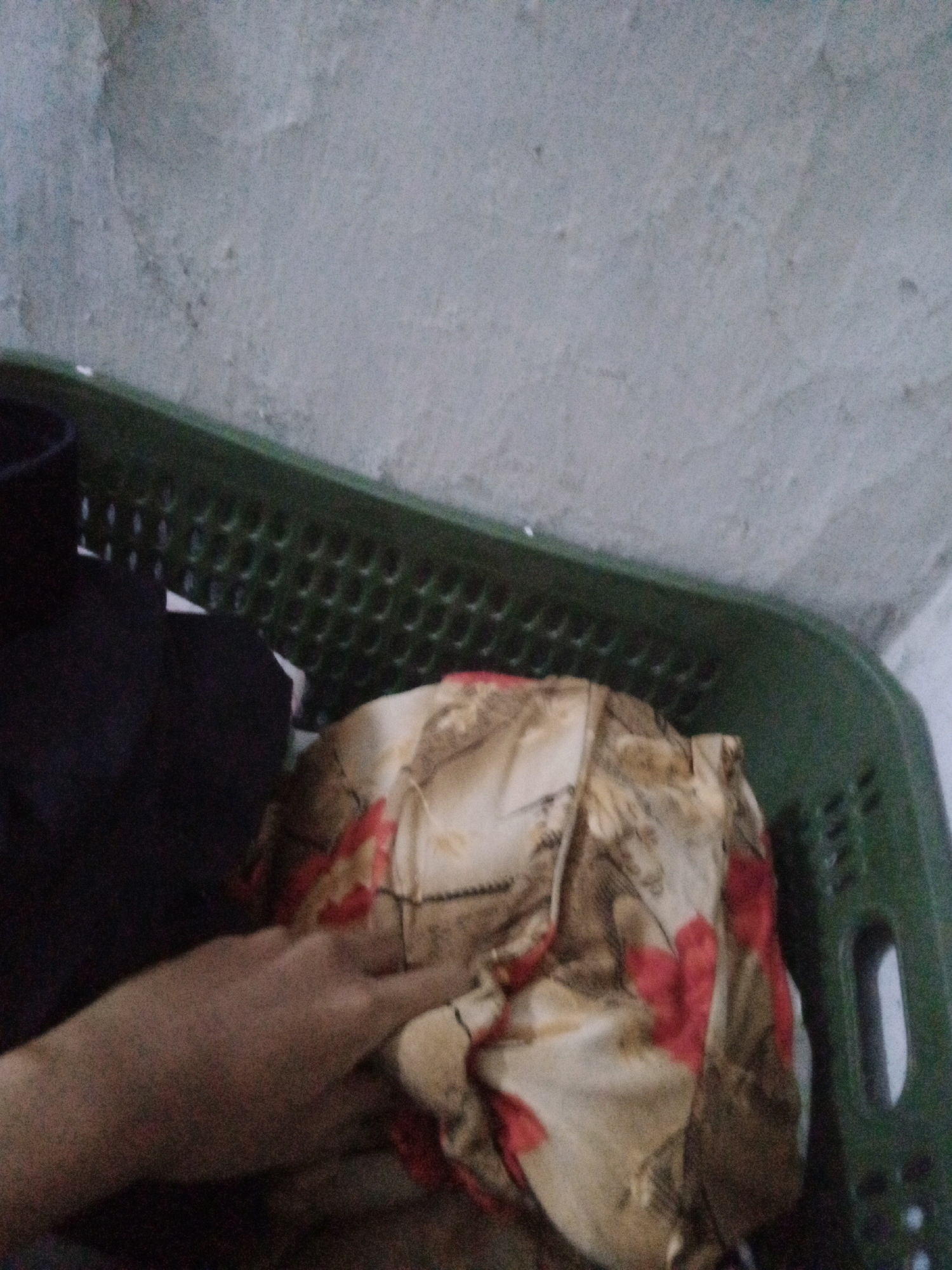 The image shows a green plastic laundry basket filled with laundry, including a patterned fabric with red and tan designs. A person's hand is visible resting on the laundry. The background appears to be a textured wall. The scene is indoors, and the lighting is dim. There are no discernible elements that would indicate the location within Boyoteluk, Indonesia.