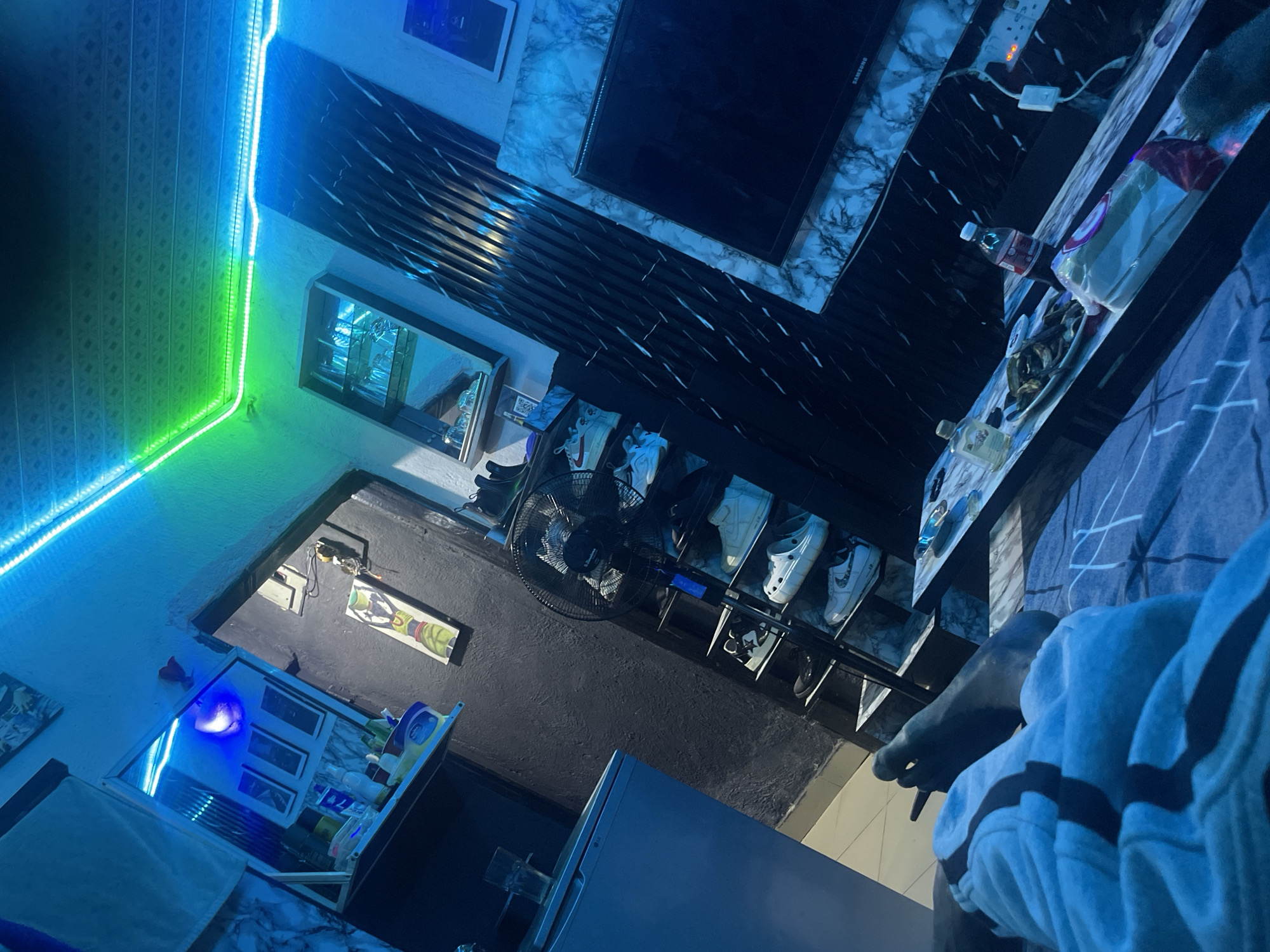 The image shows a dimly lit room with neon blue and green LED lights illuminating the space. In the foreground, a person's feet and legs are visible, covered by a light blue and white striped blanket. A coffee table sits in front of the person, with a plate of food, a bottle of soda, and other items on its surface. To the left, a tall, black fan is positioned next to a shelf unit displaying several pairs of shoes. Further to the left, a light blue refrigerator is visible. In the background, a doorway leads to another area of the room, where a picture hangs on the wall. A mirror is mounted on the wall above a shelf containing bottles. A television is mounted on the wall on the right side of the room. The overall impression is a casual, indoor setting, possibly a living room or bedroom.