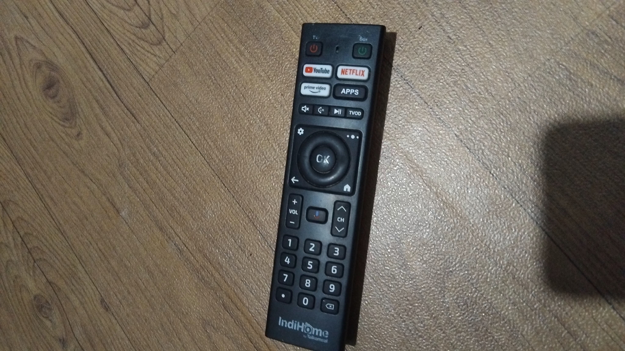 An IndiHome remote control lies on a textured, wood-grain surface, likely a floor. The remote is black and oriented horizontally, with the IndiHome logo visible on the left side. Buttons for numbers 0 through 9, as well as power, volume, channel selection, and directional navigation are present. Additionally, there are dedicated buttons for apps such as YouTube, Netflix, and Prime Video. The scene is static, with no discernible action or interaction. The environment appears to be an indoor setting, with the wood-grain pattern suggesting a floor or tabletop. No people or specific urban features of Bangun Rejo, Indonesia are visible in this image.