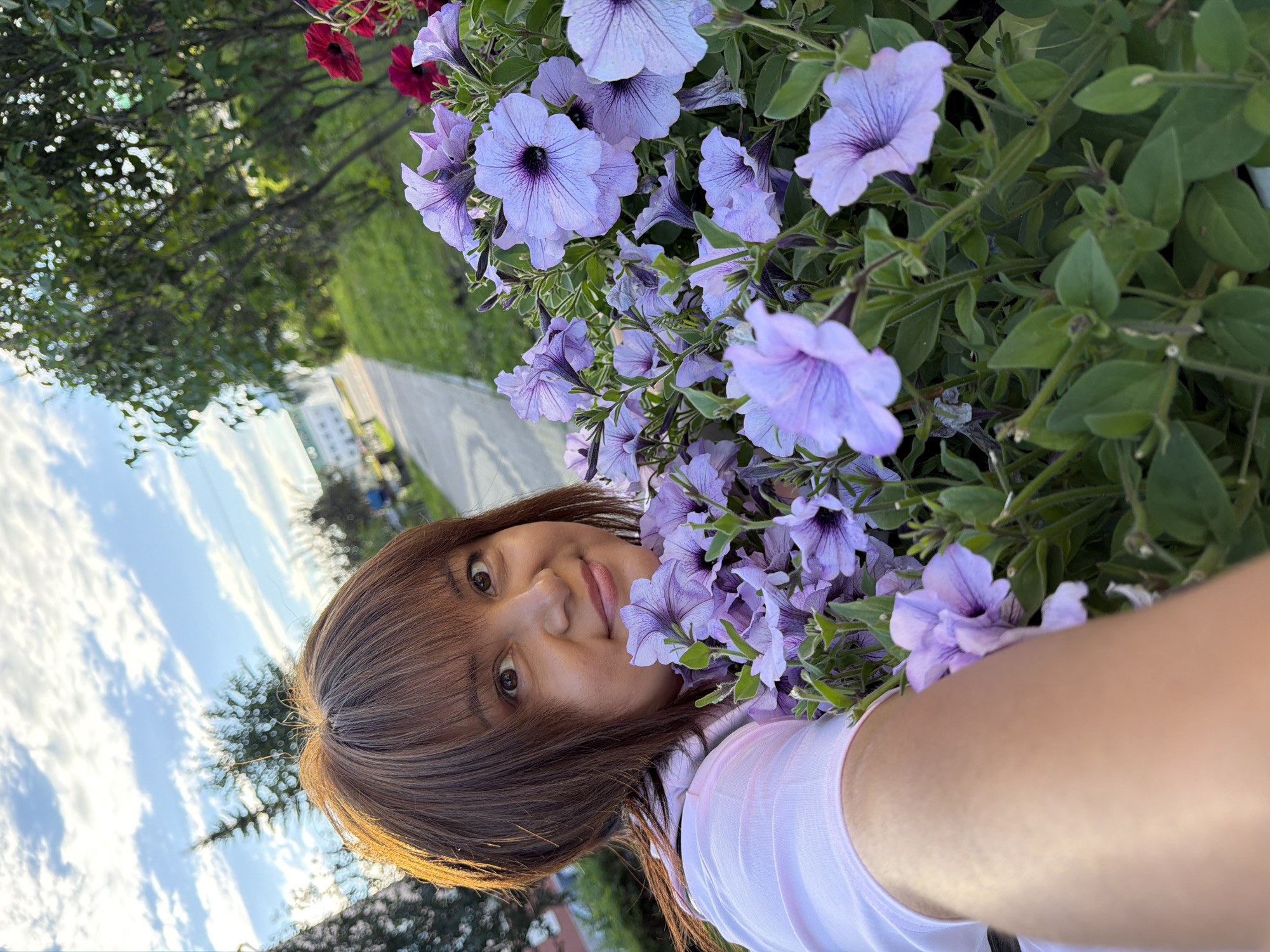 This is a selfie of a young woman with brown hair and bangs, smiling as she holds a bouquet of light purple petunias. She is wearing a light pink shirt. The background is blurred, but appears to be an outdoor setting with trees and a paved path. The sun is shining, casting a warm glow on her face and hair, suggesting it is either morning or late afternoon. The sky is visible in the upper left corner with white clouds against a bright blue backdrop. The overall mood is cheerful and relaxed.
