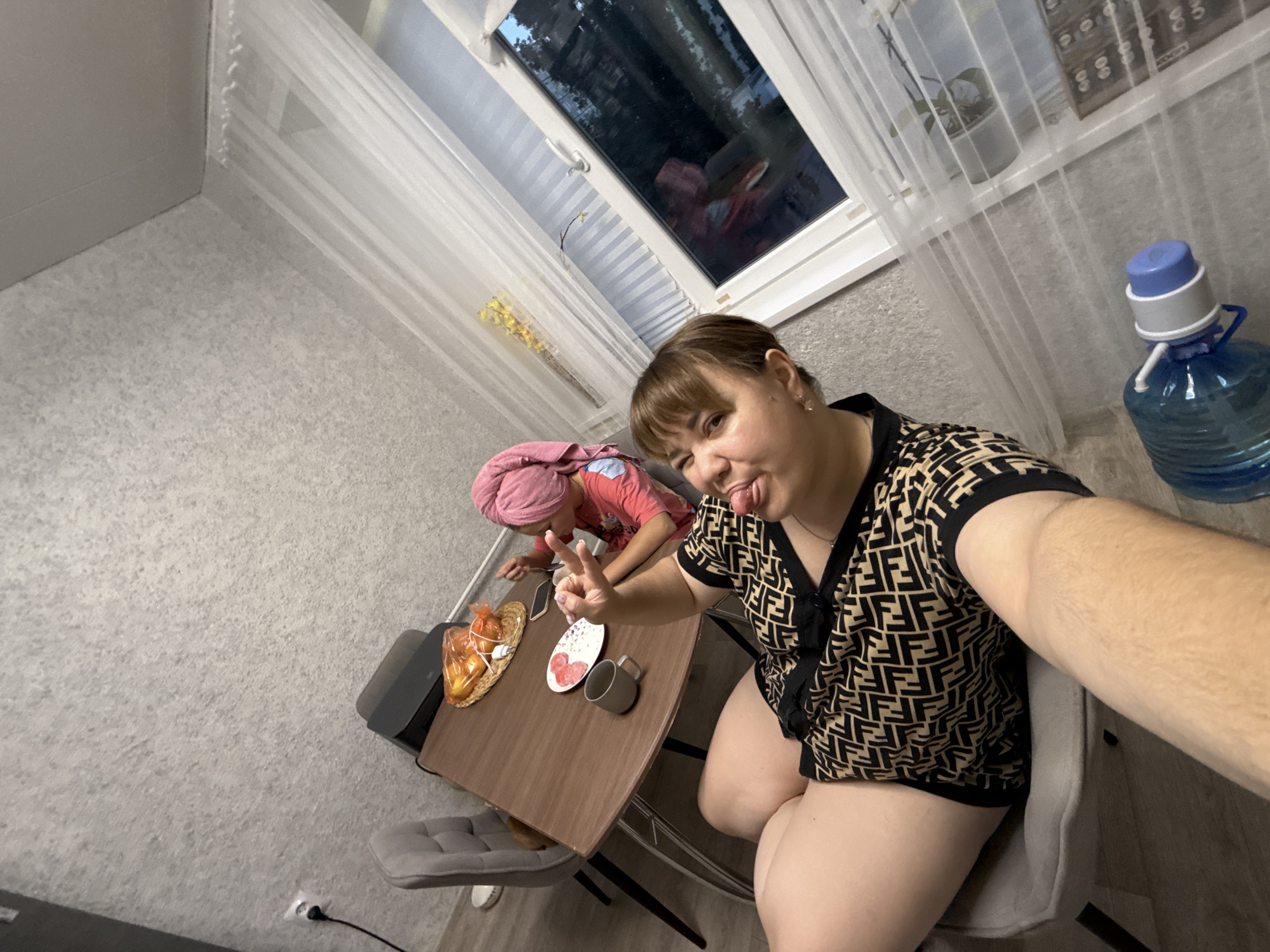 This media file is a selfie taken indoors. The main subject is a woman with brown hair, wearing a black and brown patterned shirt and black shorts. She is sticking her tongue out and making a peace sign with her fingers. She is seated in a chair facing the camera, with her legs visible.
In the background, another person, likely a child, is seated at a table wearing a pink shirt and a pink towel wrapped around their head. The table has a basket of fruit, a plate with heart-shaped candies, and a mug on it. A partially visible chair is to the left of the table.
The setting appears to be a home, with a window in the background covered by sheer white curtains. Through the window, it appears to be evening or night, with dark trees visible outside. A large blue water dispenser with a pump is located to the right of the frame. The walls have a textured wallpaper. The lighting suggests it's indoors, likely in the evening. The overall mood seems playful and casual.
