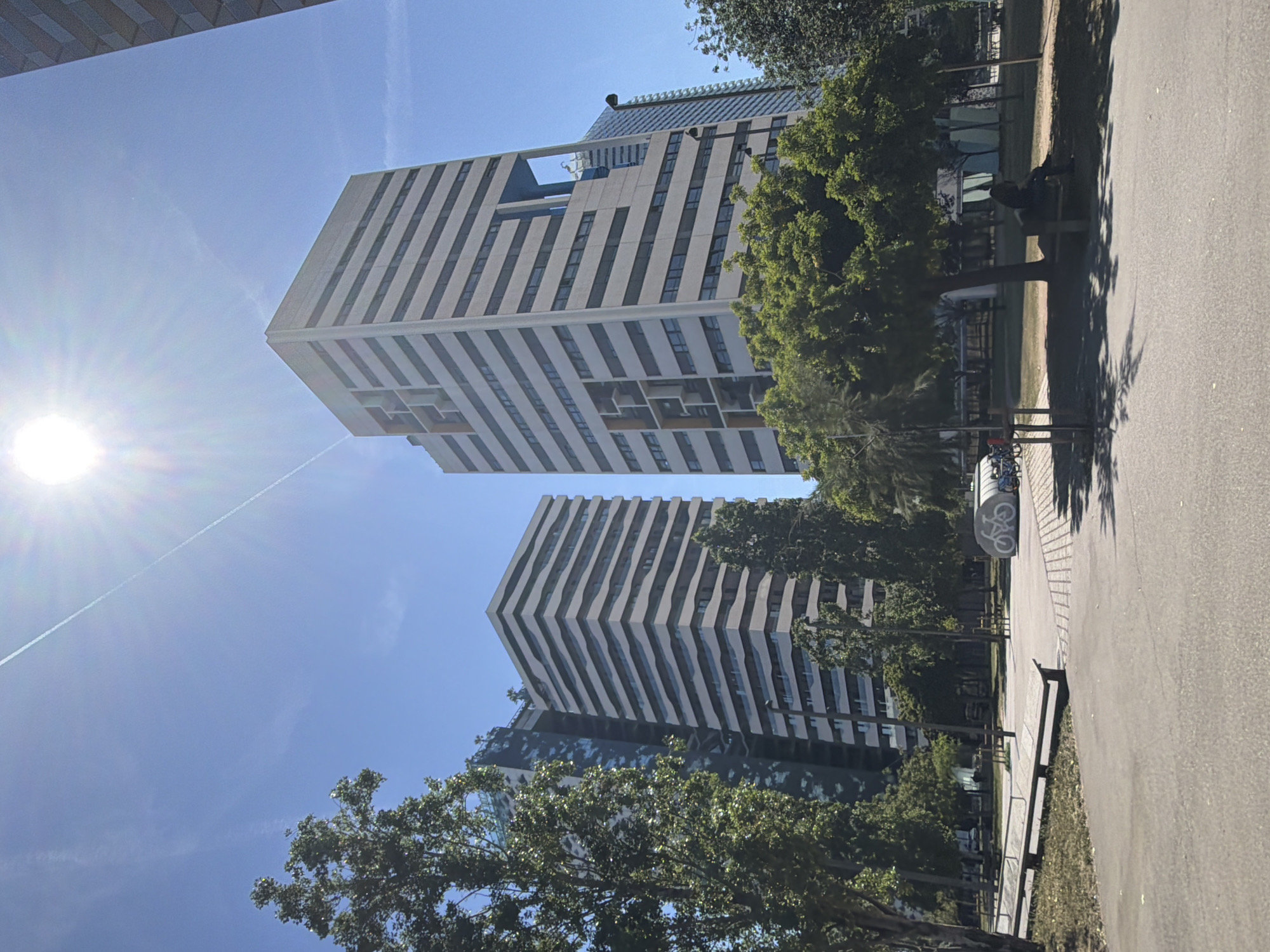 The image depicts a sunny day in the Parc de la Remunta, located in l'Hospitalet de Llobregat, Spain. Two tall, modern buildings with a striped facade dominate the background, partially obscured by trees. A paved path runs along the right side of the image, with benches and trees lining the park area. A person is sitting on one of the benches. A sign with a bicycle symbol is visible along the pathway. The sky is clear and blue with some airplane contrails. The sun is bright in the upper-left corner.