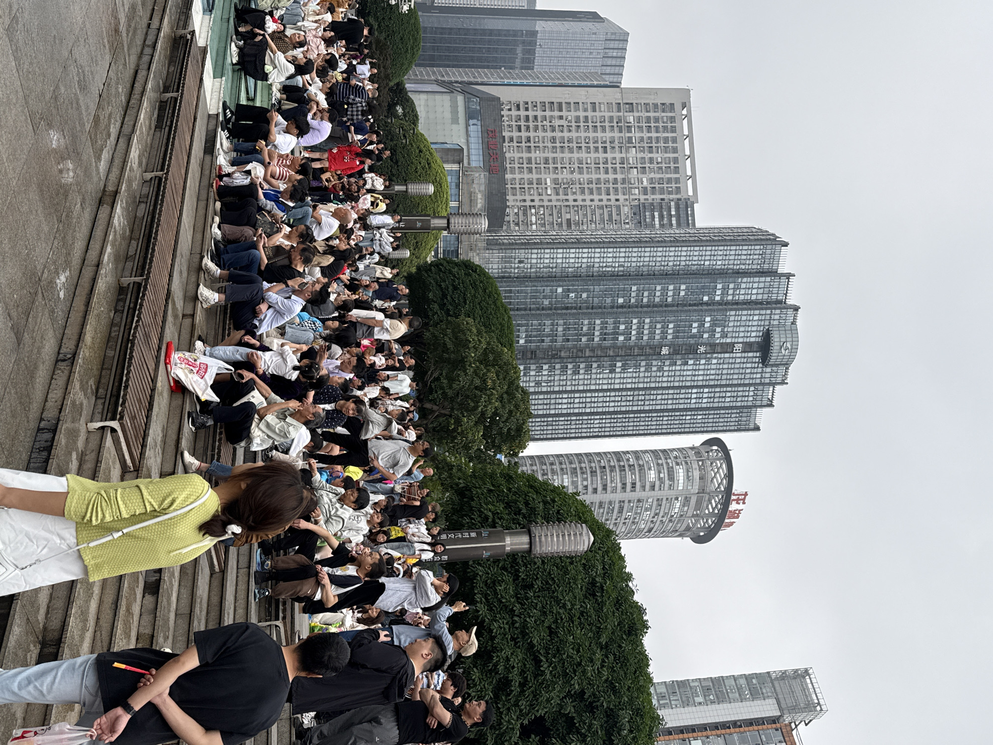 A large group of people are sitting on concrete steps in what appears to be an outdoor public space in Liangjiang, China. The people are mostly young adults, and they are casually dressed. Many are using their phones.
Behind the group are dense green bushes that separate the seating area from the cityscape. Several modern high-rise buildings of varying heights are visible in the background. The sky is overcast.
A woman in a yellow sweater and white pants is walking in the foreground. Several people in black shirts are also in the foreground, walking in the opposite direction.