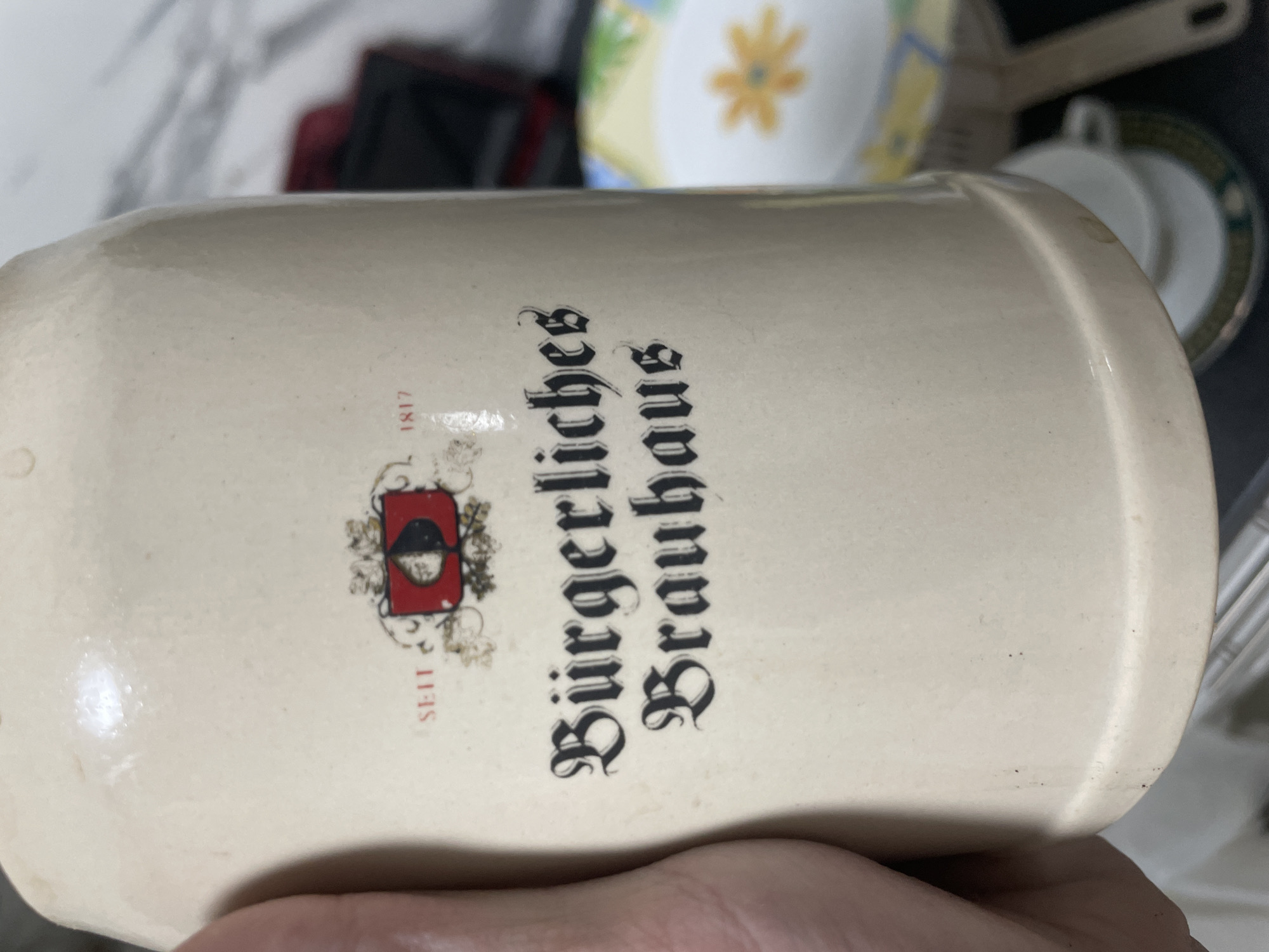 The image shows a close-up of a ceramic stein with a logo and text. The logo is a shield with a half black and half white spade symbol on a red background, framed by decorative elements. The text below the logo reads "Bürgerliches Brauhaus" in a gothic font. Above the logo, the word "SEIT" is visible on the left, and the number "1817" is visible on the right, both in red text. A hand is partially visible on the left side of the frame, holding the stein. The background is out of focus and appears to be an indoor setting with some objects that resemble dishes or other household items. There is no indication of the location being the city of Bol'shoe Zaborod'e, Russia.