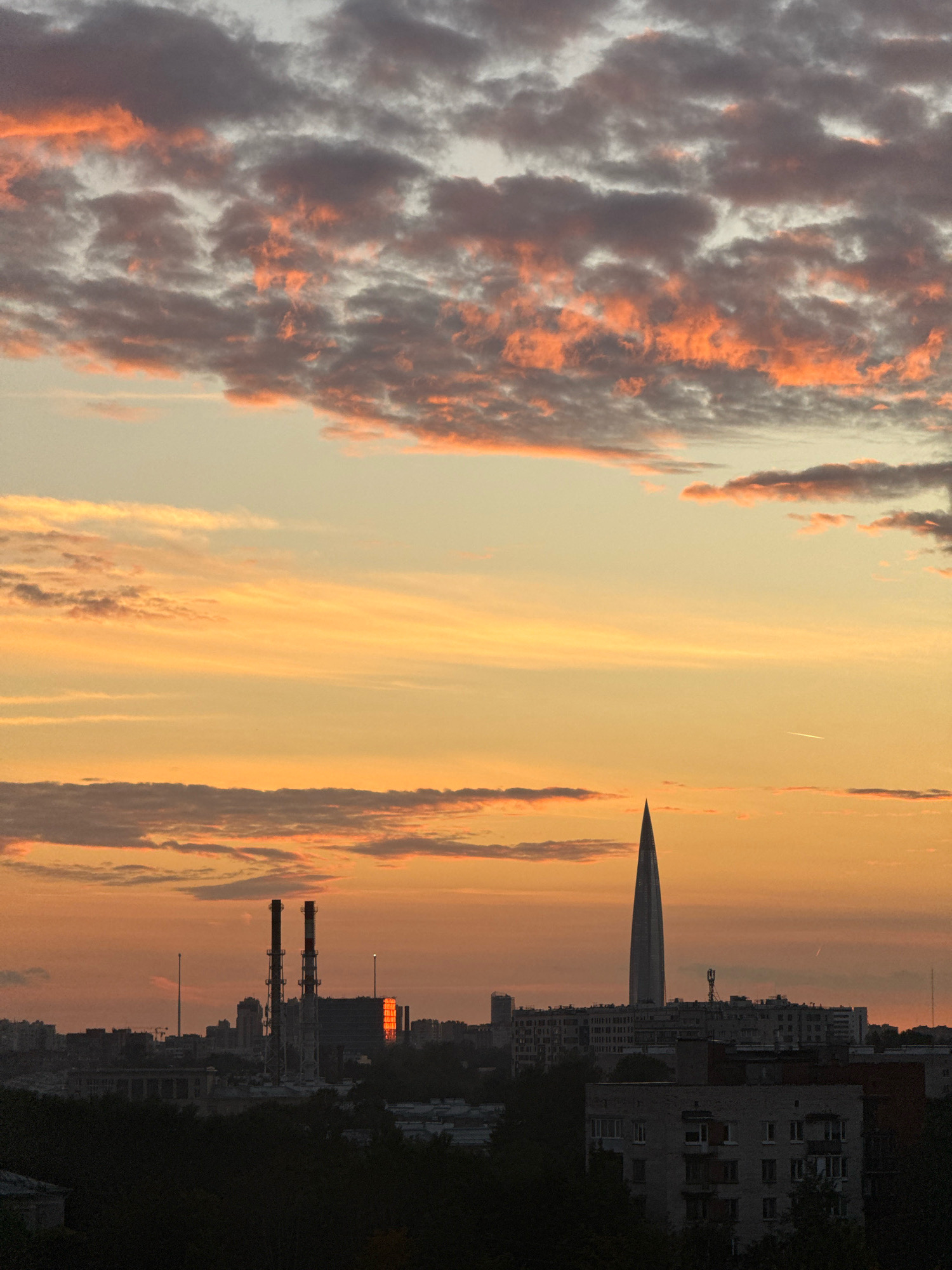 This is a photograph of a sunset over the city of Saint Petersburg, Russia. The sky is filled with dramatic clouds, tinged with orange and pink hues. In the foreground, silhouetted trees partially obscure the view of the city. Prominent in the middle ground are industrial smokestacks, with red and white banding. A tall, slender skyscraper, characteristic of modern architecture, pierces the sky in the background. The cityscape consists of various buildings, some appearing to be residential blocks, while others might be industrial or commercial structures. The overall impression is one of an urban landscape at dusk, with a mix of industrial and modern architectural elements under a vibrant sky.