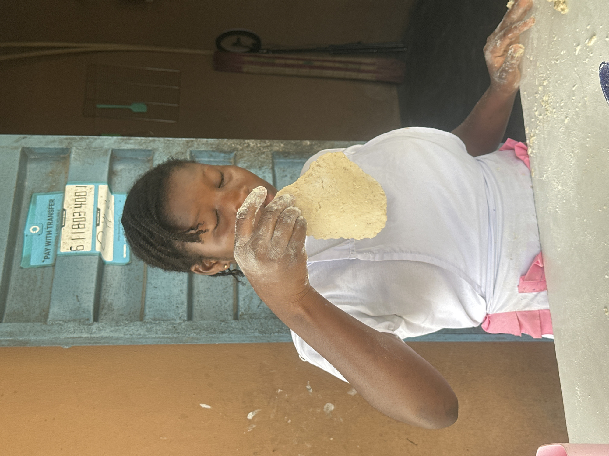 A young woman with dark skin and braided hair, wearing a white t-shirt and a pink and white apron, is seen in what appears to be a shop or eatery in Badagry, Nigeria. She is holding a flat piece of dough in her hands, and her fingers are dusted with flour. The background features a blue corrugated metal door with a sign that says "PAY WITH TRANSFER" and a phone number. To the right, there is a dark doorway with a rack of items and a circular object that might be a fan or wheel. The woman's eyes are closed, suggesting concentration as she works with the dough. The overall environment appears to be functional and perhaps a bit worn, consistent with many businesses in the area.