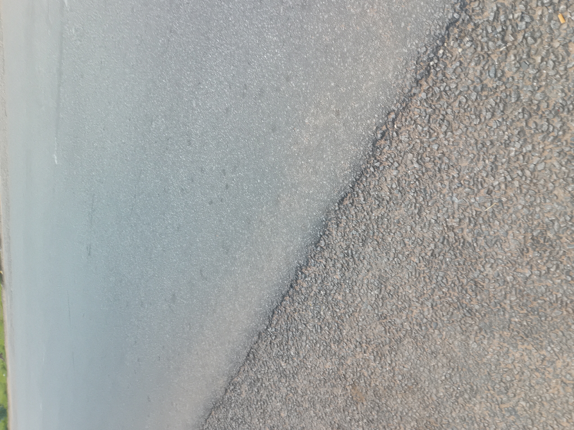 This is a ground-level, eye-level, medium shot image of a paved road. The road appears to be a two-lane highway, with the left lane appearing to be a rougher, more gravelly asphalt, and the right lane a smoother, finer asphalt. The dividing line between the two lanes is a diagonal line starting from the lower left corner of the image and extending towards the upper right. The road is in good condition, with no visible cracks or potholes. The environment appears to be an outdoor, daytime scene. There are no people or vehicles visible in the image. The location is likely a rural area, as there is vegetation visible in the background on the left side of the image. The image may have been taken in Jalingo, Nigeria, a city in northeastern Nigeria known for its large agricultural sector and arid climate.