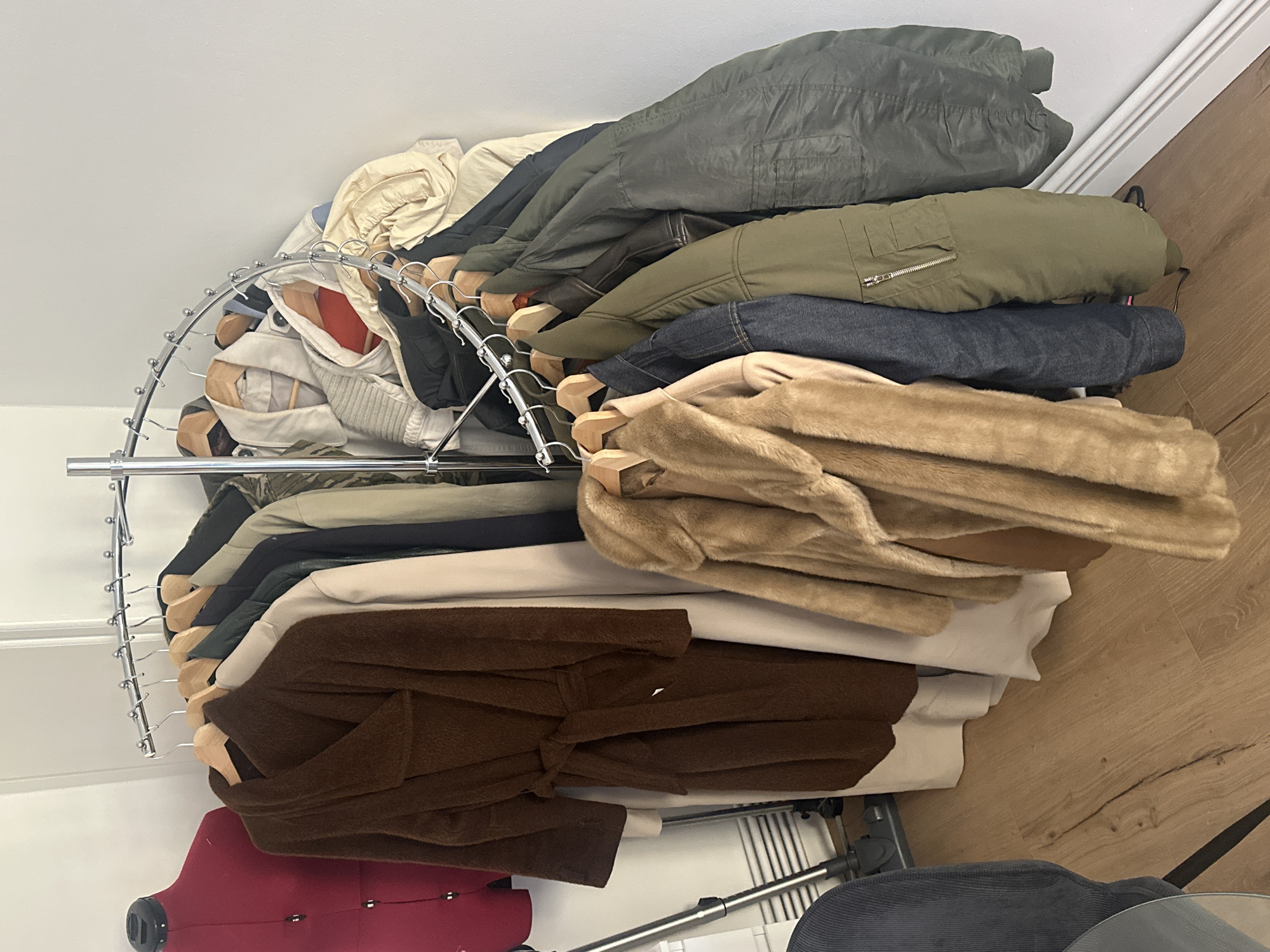 The image shows a cluttered, indoor scene with a circular clothing rack overloaded with various garments. The rack is silver and chrome, displaying a range of coats, jackets, and a brown robe. The clothing is piled high, suggesting limited space or organization.
To the left of the rack is a red dress form mannequin. The background includes a white wall and a section of light brown wood flooring. The scene is likely within a private residence or studio, but there are no clues to suggest this is in London, United Kingdom, or any other city.