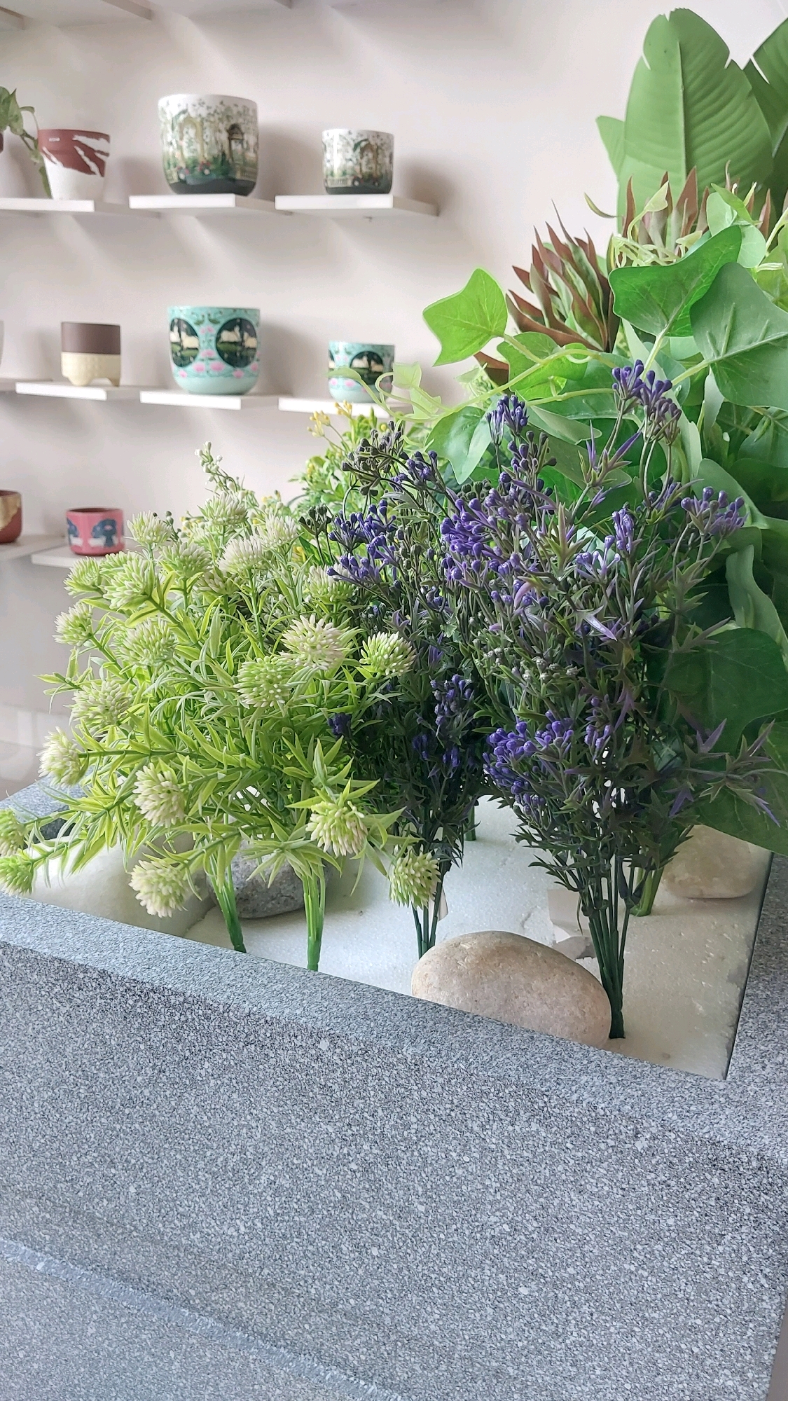 The image depicts a close-up view of a planter filled with artificial plants. Two main types of artificial plants are visible: one with light green, spiky foliage and small white flower-like clusters, and another with dark green stems and small purple buds. Interspersed with these are larger, artificial green leaves, possibly from a banana plant. The base of the planter appears to be filled with white material, possibly styrofoam, with a few smooth, light-colored stones placed among the plants. The planter itself is made of a textured gray material with a speckled appearance. In the background, several small decorative pots are displayed on white shelves. These pots are ceramic and feature intricate painted designs, including landscape scenes and geometric patterns. The overall scene appears to be an indoor arrangement, likely for decorative purposes. The location of this scene in Pune, India cannot be determined from the provided image.