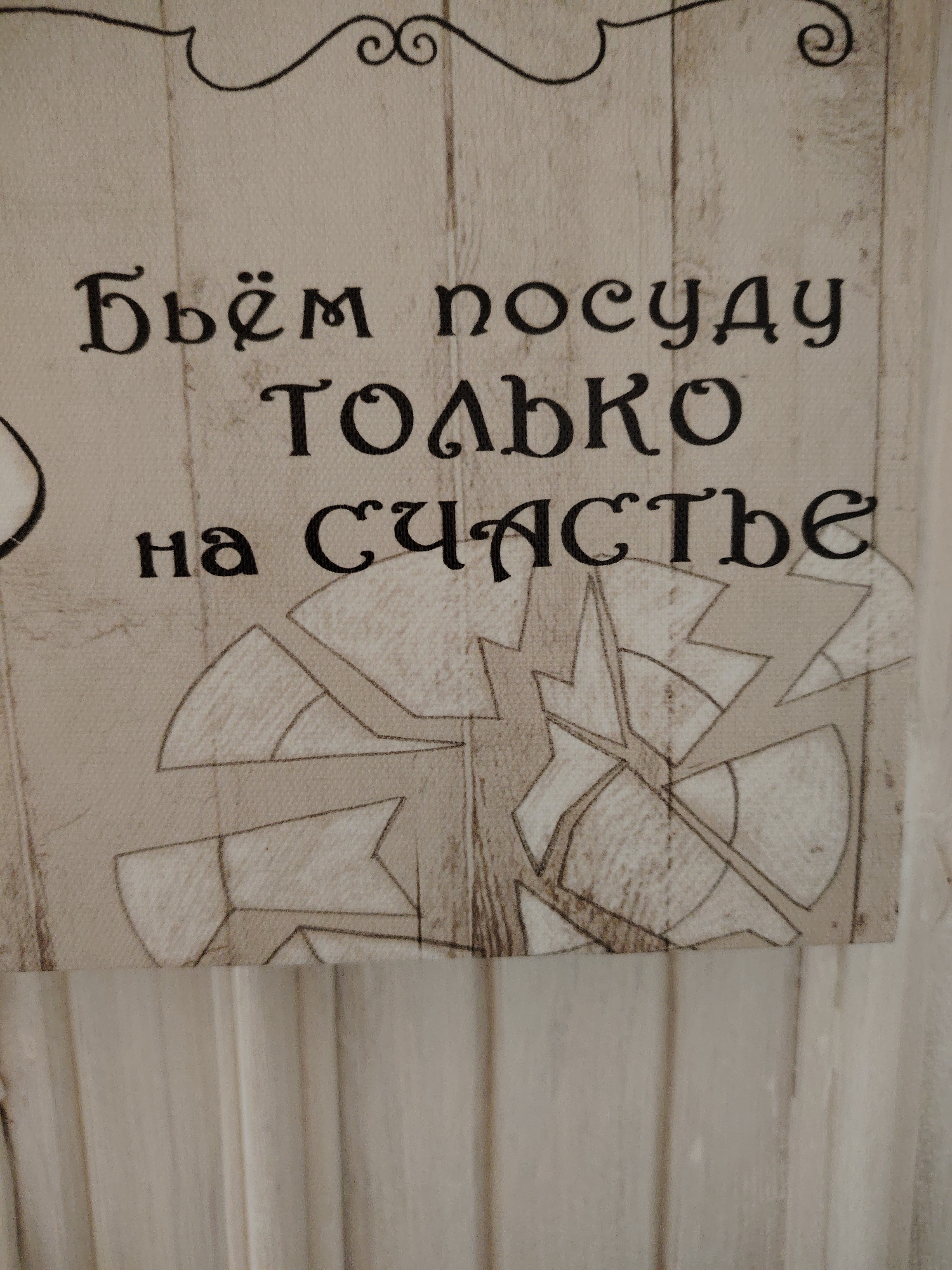 The image shows a close-up of a textured surface with Cyrillic text printed in black. The text appears to be a decorative element, possibly on a wall hanging or fabric. The words read "Бьём посуду только на счастье" which translates to "We break dishes only for good luck." Below the text, there is a stylized graphic that resembles broken pieces of pottery or glass arranged in a circular pattern. The background has a wood-grain texture. The image does not contain any elements related to the city of Vladimirovka, Russia.