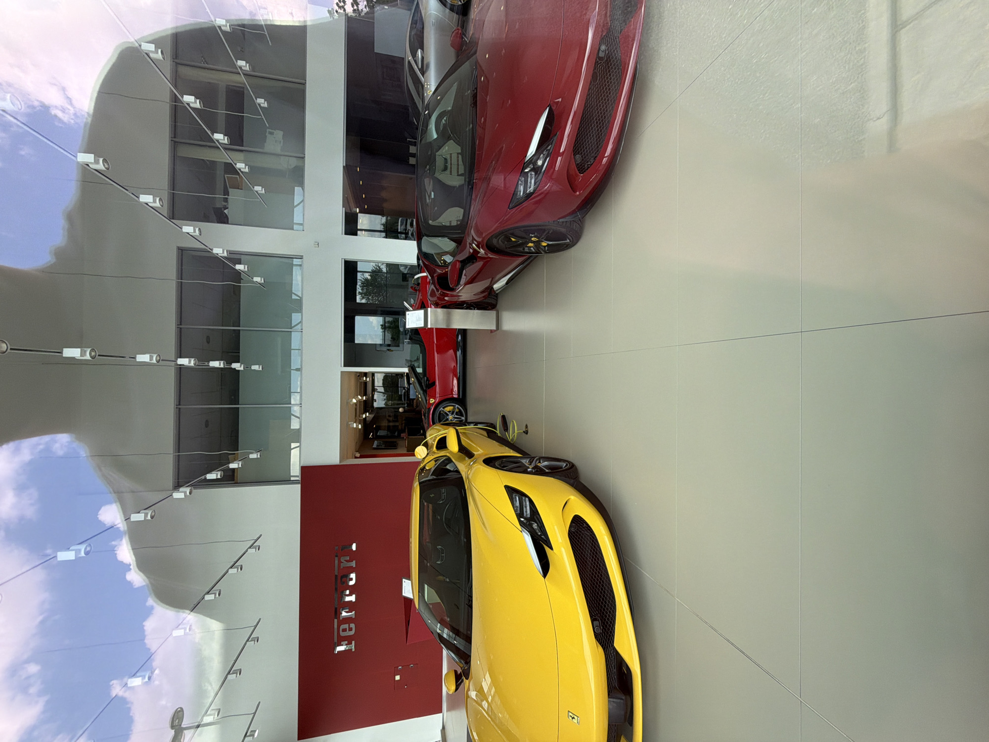 This image depicts the luxurious interior of a Ferrari showroom, likely located in Busmantsi, Bulgaria. The primary subjects are two high-performance sports cars: a bright yellow Ferrari positioned prominently in the foreground, and a red Ferrari situated further back. Both vehicles are meticulously displayed on a pristine, light-colored tiled floor, with their front fascias angled towards the right side of the frame.
The setting is characterized by modern architecture, featuring extensive glass facades that serve as the exterior walls of the showroom. These glass surfaces strongly reflect the daytime sky, showing distinct blue patches and white clouds, suggesting clear or partly cloudy weather. Reflections of internal lighting fixtures and structural elements of the building are also visible on the glass.
A notable location cue is a bold red wall on the left side of the image, clearly displaying the brand name "Ferrari" in white, three-dimensional lettering. This text unmistakably identifies the establishment. The clean lines, bright lighting, and polished surfaces contribute to an atmosphere of exclusivity and sophistication. No individuals are visible, maintaining a focus on the showcased automobiles. The scene reflects a sunny daytime setting, enhancing the vibrant colors of the cars.