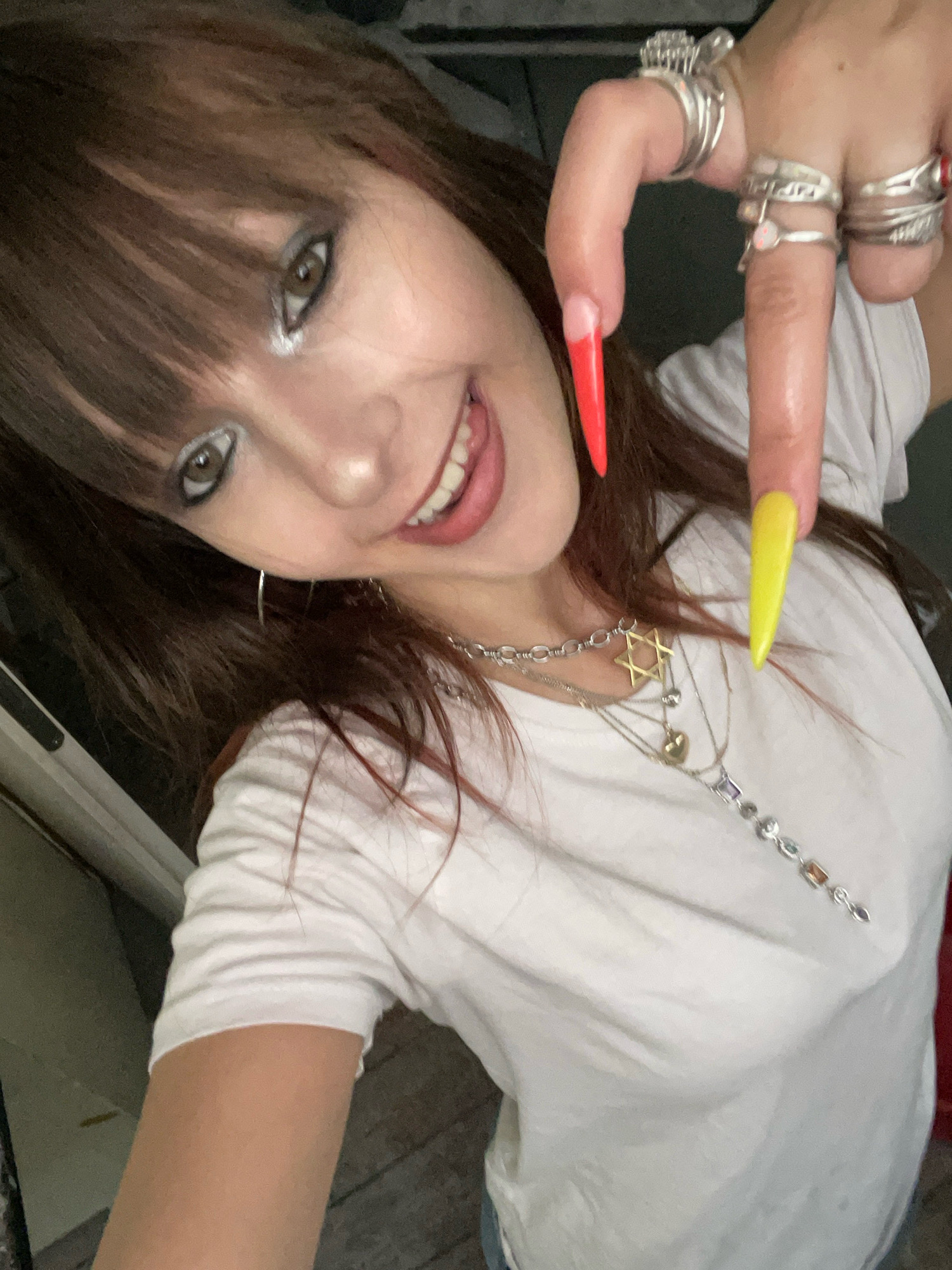 A young woman with brown hair and bangs, wearing a white t-shirt and multiple silver necklaces, poses for a selfie. She has dramatic eye makeup, including silver eyeshadow and dark eyeliner. Her nails are long and pointed, with the index finger nail painted red and the middle finger nail painted yellow. She is wearing several silver rings on her fingers. The background appears to be indoors, with hints of what looks like a door frame and wooden flooring, suggesting a residential setting. There are no discernible features that specifically place the image in Raanana, Israel.