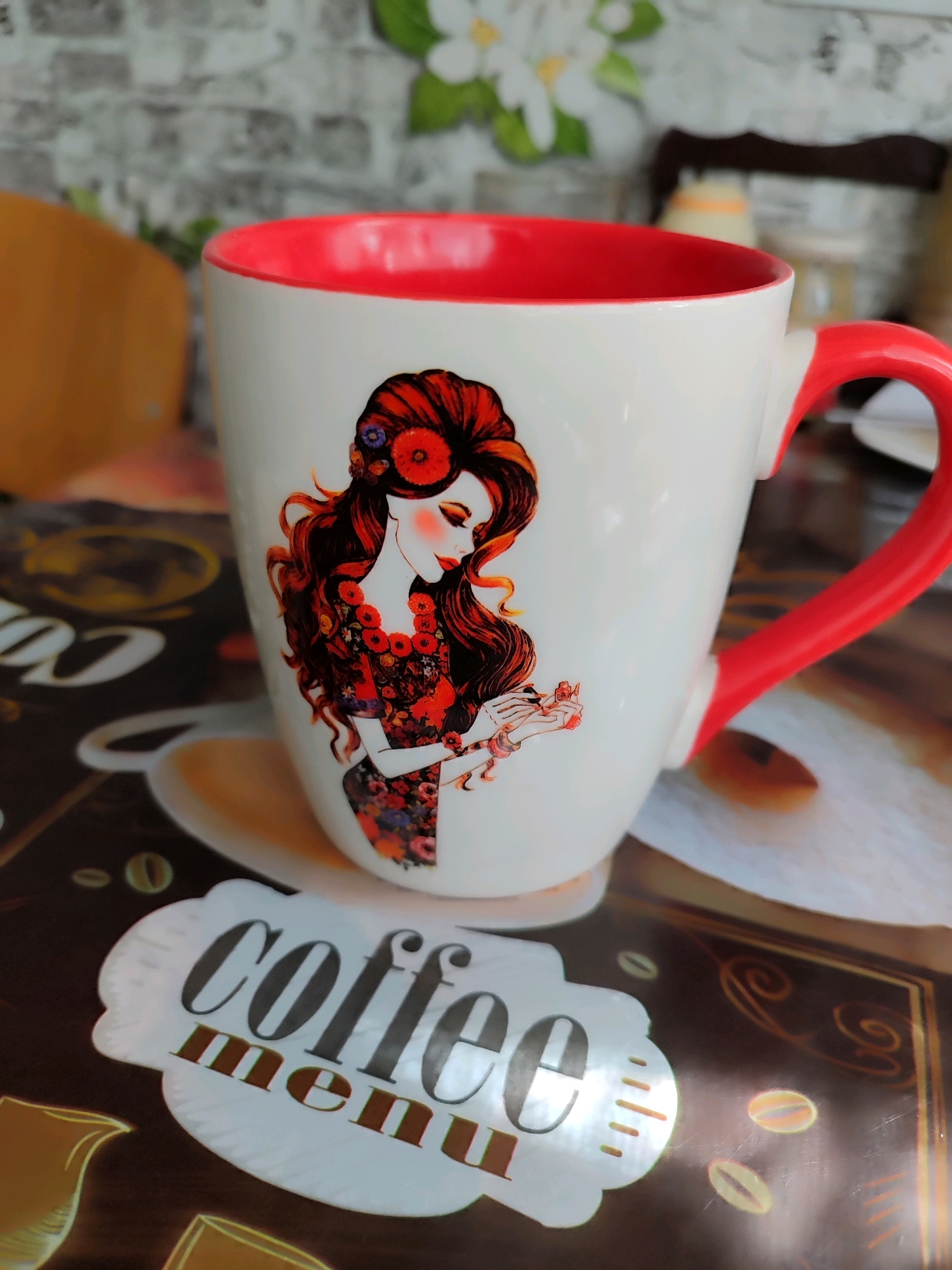 A white coffee mug with a red interior and handle is shown, decorated with a painting of a woman with flowing red hair and a floral dress. The mug is on a dark surface with a coffee-themed pattern, including coffee beans. A sticker with the words "coffee menu" is placed in front of the mug. The background is blurred and shows a brick wall and some green leaves.