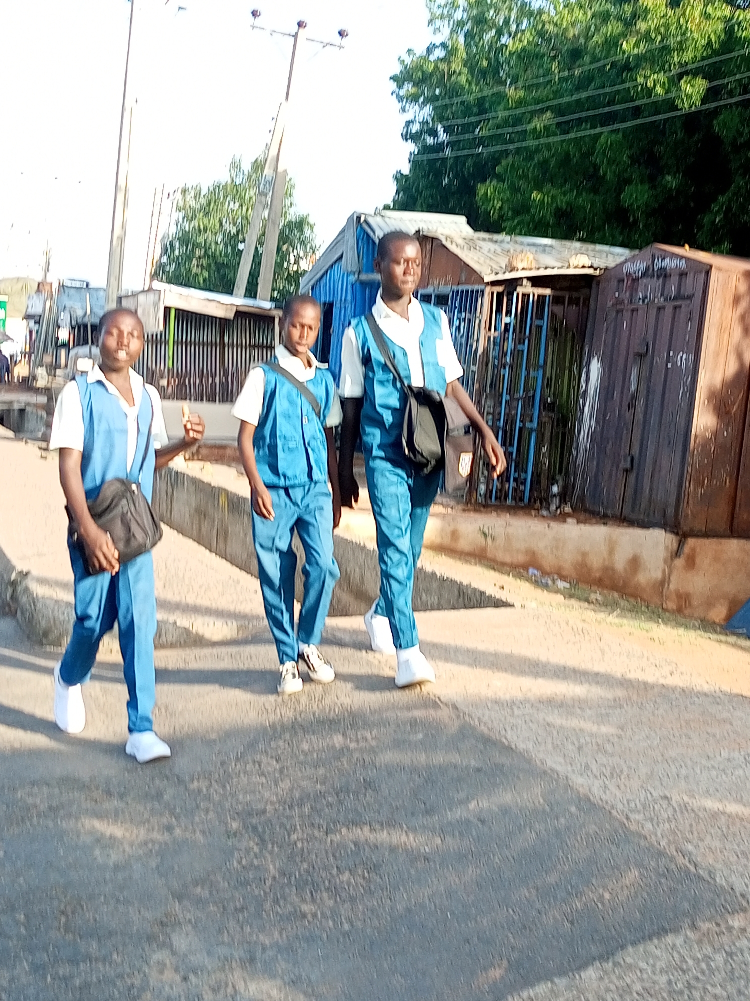 The image shows three young people in blue and white school uniforms walking on a paved surface. The person on the far left is holding a black bag. The middle person is wearing a black bag across their chest. The person on the far right is partially visible. In the background, there are structures that appear to be informal shops or stalls, with corrugated metal roofs and barred entrances. Trees are visible behind the structures, and utility poles with wires stretch across the sky. The overall environment suggests an urban or semi-urban setting in Tempure, Nigeria. The lighting indicates it is daytime, likely late afternoon due to the long shadows cast on the ground.
