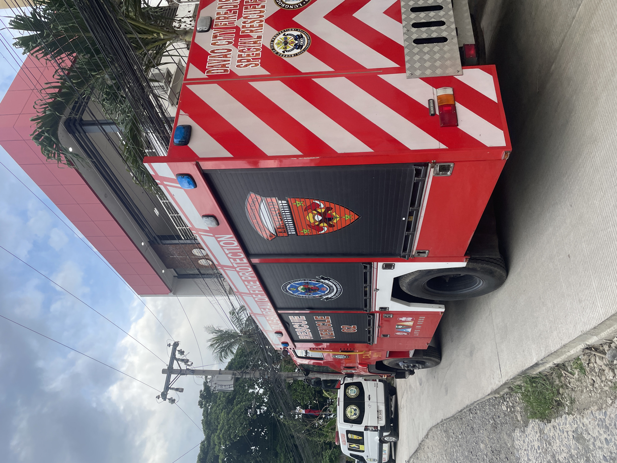 An eye-level, medium shot captures a red and white Davao City Fire District Special Rescue Vehicle parked on a concrete surface in Davao City, Philippines. The fire truck, detailed with diagonal red and white stripes, features "RESCUE VEHICLE 02" emblazoned on its side along with several official emblems. To the left, a white emergency van with similar emblems and the license plate "LAJ 3456" is partially visible. The background includes a modern building with a red facade, electrical poles, wires, lush green trees, and a cloudy sky. The scene appears to be on a street or pathway adjacent to a building.