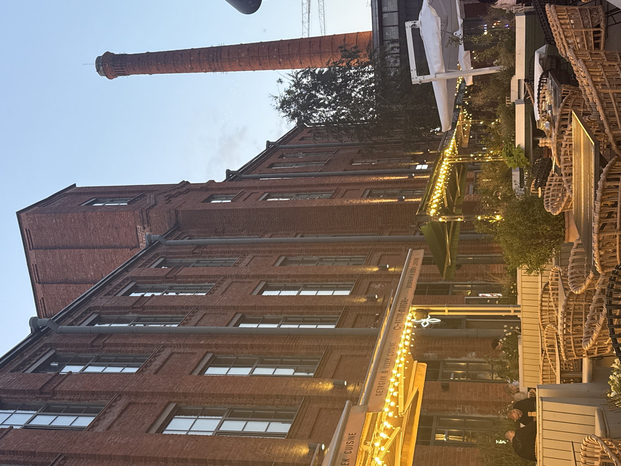 The image shows an outdoor seating area of a restaurant in Saint Petersburg, Russia, likely located in an industrial or converted factory area. The restaurant is situated next to a tall, red brick building with multiple windows. A tall, red brick chimney stands in the background, a characteristic feature of former industrial sites in Saint Petersburg, such as the L'Etoile complex or the Sevkabel Port.
The outdoor seating area features numerous wicker chairs and tables, some covered by yellow and green awnings. String lights are draped across the awnings, creating a warm ambiance. Several plants are visible in planters around the seating area.
In the foreground, there are several tables set with salt and pepper shakers and plates. Two people are seated at one of the tables, engaged in conversation or eating. Other patrons may be present but are not clearly visible. The lighting suggests it is either dusk or evening. The overall scene depicts a dining establishment in a historically industrial part of Saint Petersburg, with a blend of old architecture and modern outdoor dining.