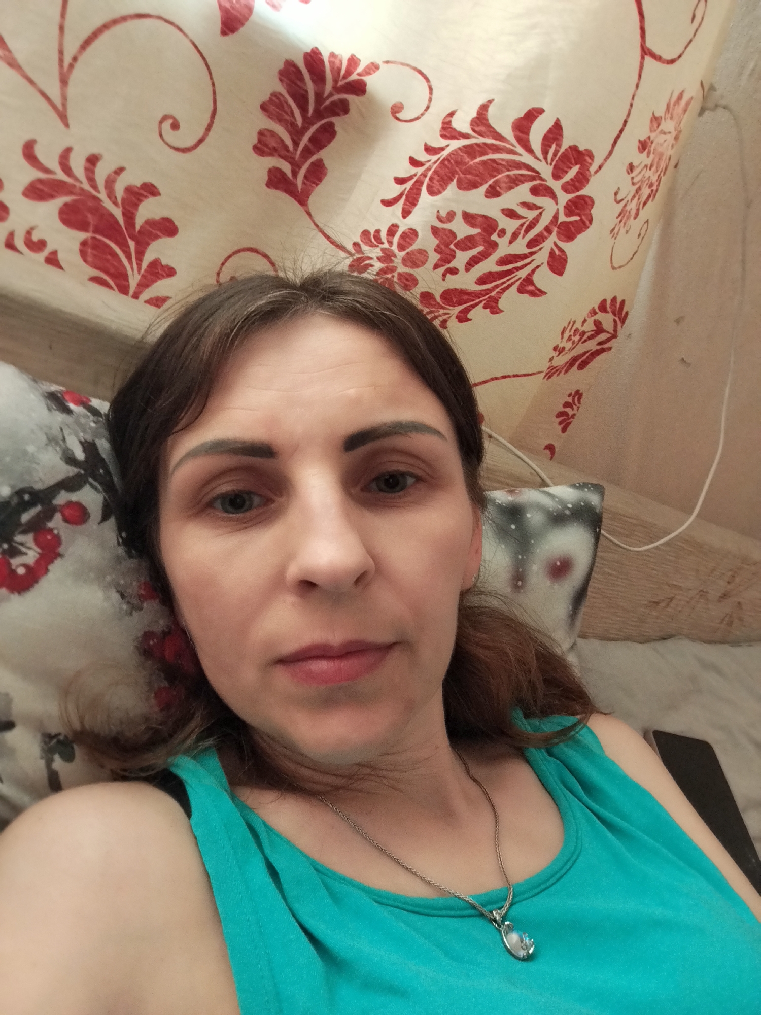 A woman is lying on a bed, looking at the camera. She has brown hair and is wearing a turquoise tank top and a silver necklace. The background consists of pillows and a patterned curtain. The image was taken inside a residential dwelling in Vinnytsia, Ukraine.