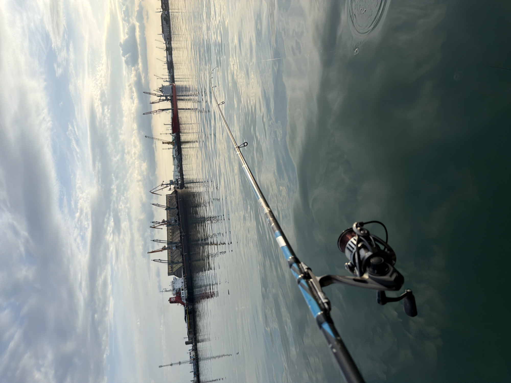The image shows a fishing rod in the foreground, with a harbor scene in the background. The water is calm, reflecting the sky and the buildings in the harbor. Cranes, ships, and other port infrastructure are visible on the shore. The fishing rod is angled towards the water, suggesting someone is actively fishing. Based on the visible characteristics and the style of the image, the location is consistent with the harbor area in Burgas, Bulgaria, specifically in the port area near the industrial structures and cranes. The fishing rod and reel are in the right of the image. The hook and line is visible in the water above the fishing rod.