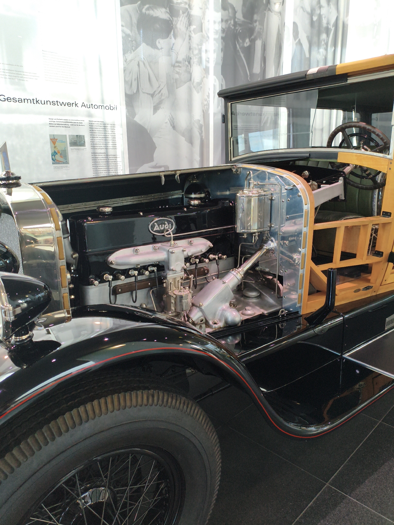 The image shows a close-up view of the engine compartment and front section of a vintage Audi automobile. The engine is black with silver and chrome accents, and the Audi logo is prominently displayed on the engine block. The car has a wooden interior frame visible behind the engine. The exterior is black with a red pinstripe along the fender. A spoked wheel with a ribbed tire is visible on the lower left. In the background, a large black and white photograph or banner depicts a crowd of people. Text is visible in the upper left corner of the image, with the words "Gesamtkunstwerk Automobil" clearly legible. This automobile is likely on display at the Audi Forum in Ingolstadt, Germany, a museum dedicated to the history of the Audi brand. There are no people visible in the immediate foreground or interacting with the car.