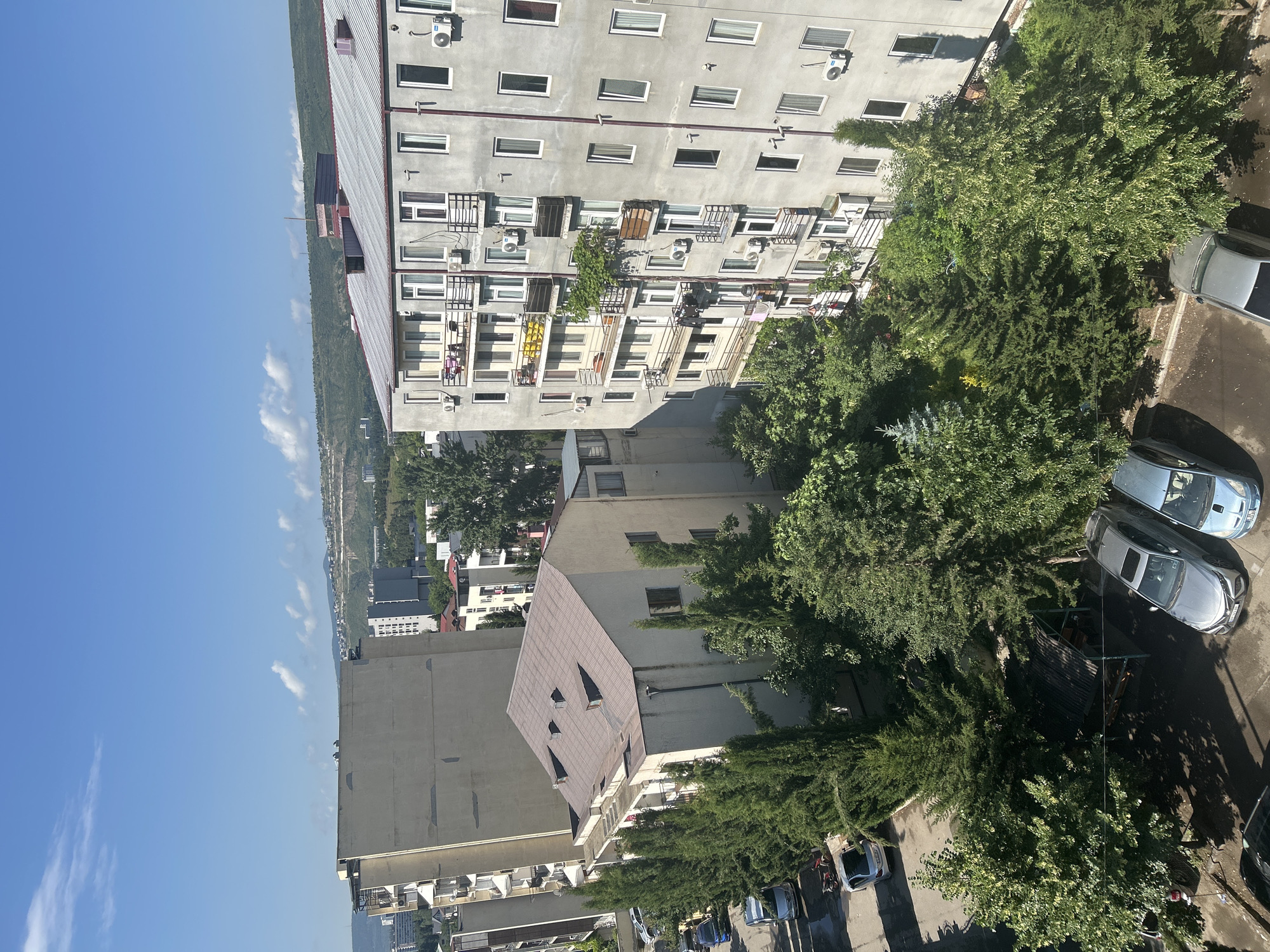 The image displays a view of residential buildings and a street in an urban setting. The buildings are multi-story, with a mix of architectural styles. One prominent building on the right is a grey, multi-story apartment complex with numerous windows and balconies. Another building to the left is a lighter-colored, taller structure. In the foreground, several cars are parked on a paved street, and a line of trees runs along the side of the road. The sky is blue with scattered white clouds. The scene appears to be captured during daylight hours, under clear weather conditions.