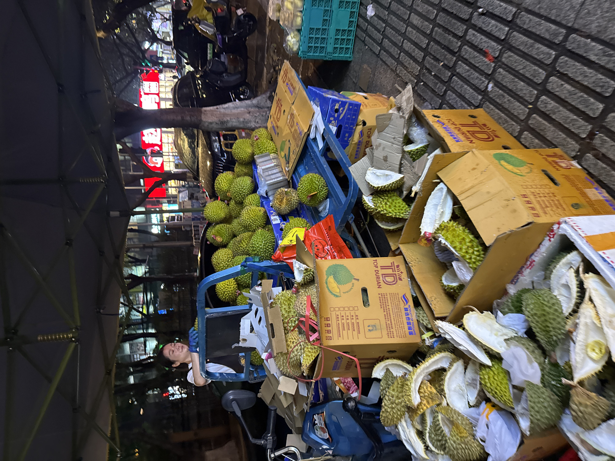 The image depicts a street scene, likely a market area, in the Tianhe District, China. A person, possibly a vendor, is sitting on a blue vehicle that appears to be a modified electric scooter or cart. The vehicle and surrounding area are piled high with durian fruits, both whole and in discarded rinds. Boxes labeled "Top Durian" are present, indicating that the vendor is selling durians. The background features a brick wall, other parked vehicles, and a glimpse of urban structures with red neon signage, characteristic of Chinese street signage at night. The scene suggests a bustling urban marketplace or impromptu fruit stall.