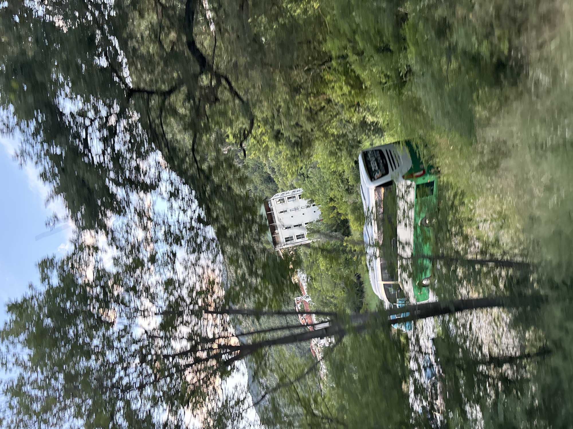 The image depicts a verdant, mountainous landscape in Petrovac centar, Montenegro, characterized by significant motion blur, suggesting it was captured from a moving vantage point. The foreground is composed of heavily blurred green foliage, including tree trunks and leaves, appearing as horizontal streaks. In the midground, a multi-story white building with a dark-edged roof and multiple dark window openings stands prominently on a hillside, surrounded by dense trees. Further left on the slope, other structures with red-tiled roofs are partially visible amidst the vegetation. At a lower elevation, a white and green passenger bus, marked with "ZENITH" and "TOUR" (or "TOURS") on its rear, is positioned on a light-colored, possibly rocky or sandy surface. A second, lighter-colored vehicle is partially visible behind it. The background consists of steep, forested mountains under a sky displaying patches of light blue and streaked white clouds, some with an orange tint, indicative of late afternoon or early morning light. No human figures are identifiable within the scene.