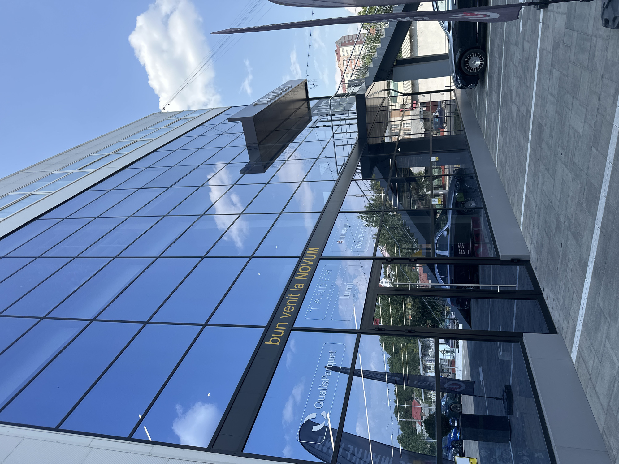The image shows the exterior of a modern, multi-story building with a large glass facade in Chișinău, Moldova. The building is branded with the name "NOVUM" and displays signage for various businesses, including "QualisParquet," "TANDEM," and "lumi." The facade reflects a clear blue sky with some clouds. In front of the building, there is a paved parking lot with several parked cars, including a dark luxury sedan and a blue sedan. An outdoor staircase with metal railings leads to an upper level of the building. The overall scene suggests a commercial or office complex.