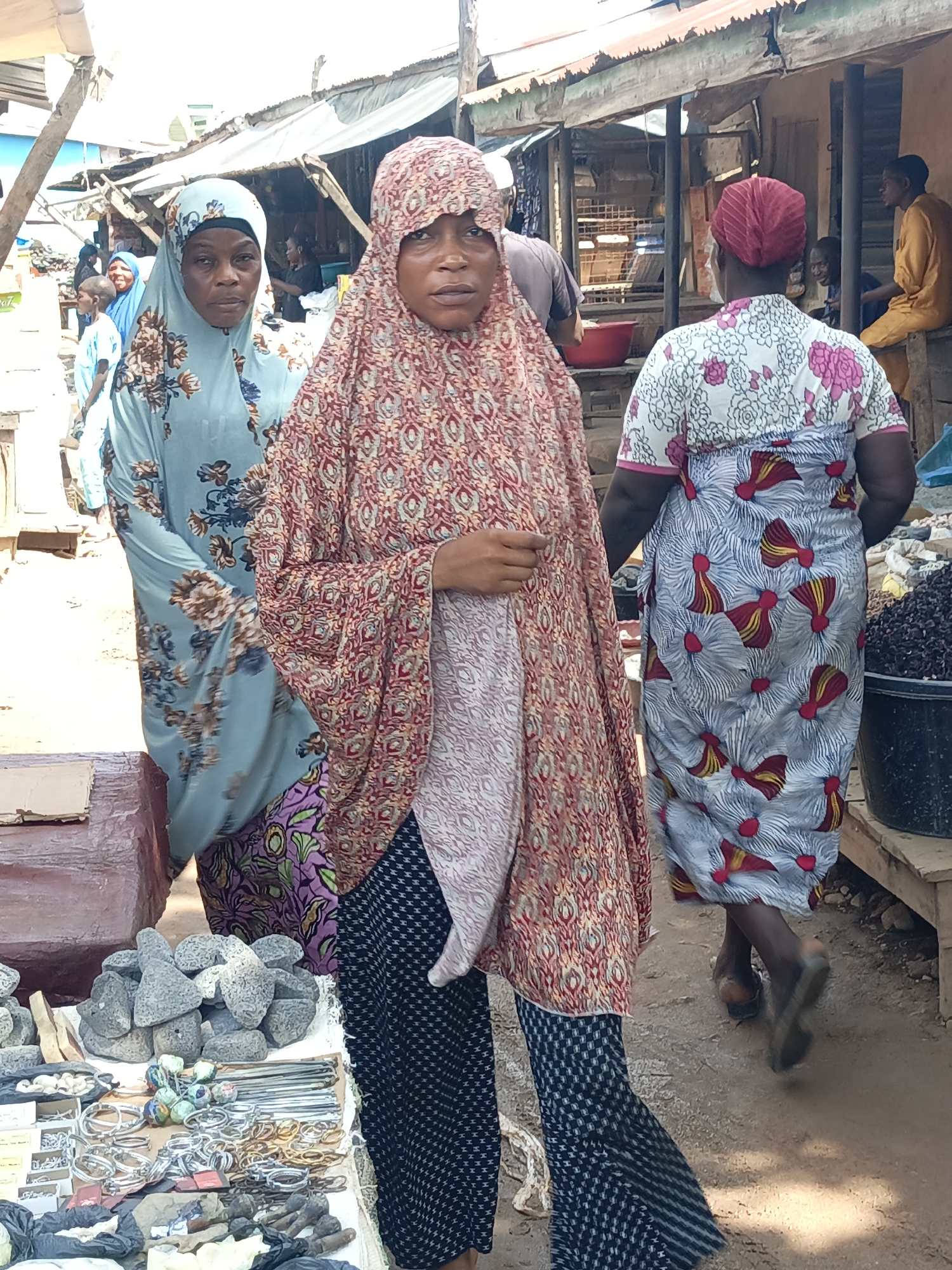 Akwanga, Nigeria. An open-air market scene shows three women in traditional clothing. The woman in the foreground wears a red patterned headscarf and a floor-length gown over black and white patterned pants. She is walking towards the viewer. To her left, another woman wears a pale blue floral headscarf and gown. To the right, the back of another woman is visible, wearing a red head covering and a patterned dress. In the background, market stalls are visible, with vendors and customers present. In the foreground, a table displays stones, bracelets, and other items for sale.