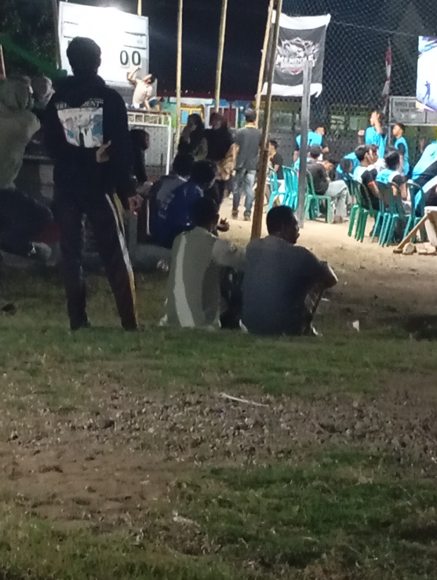 In Pacitan, Indonesia, this nighttime image captures a gathering of people, likely at an outdoor event. In the foreground, two men are seated on the grass, facing away from the camera. One man wears a dark t-shirt and pants, and the other wears a grey t-shirt and pants. Behind them, a group of individuals are visible, some standing and others sitting on green chairs. A scoreboard displaying "00" is prominent in the background, suggesting a sporting event or competition. A large black banner with white lettering hangs from a pole, and a chain-link fence forms a boundary behind the spectators. The ground is a mixture of dirt and grass, and artificial lighting illuminates the scene.