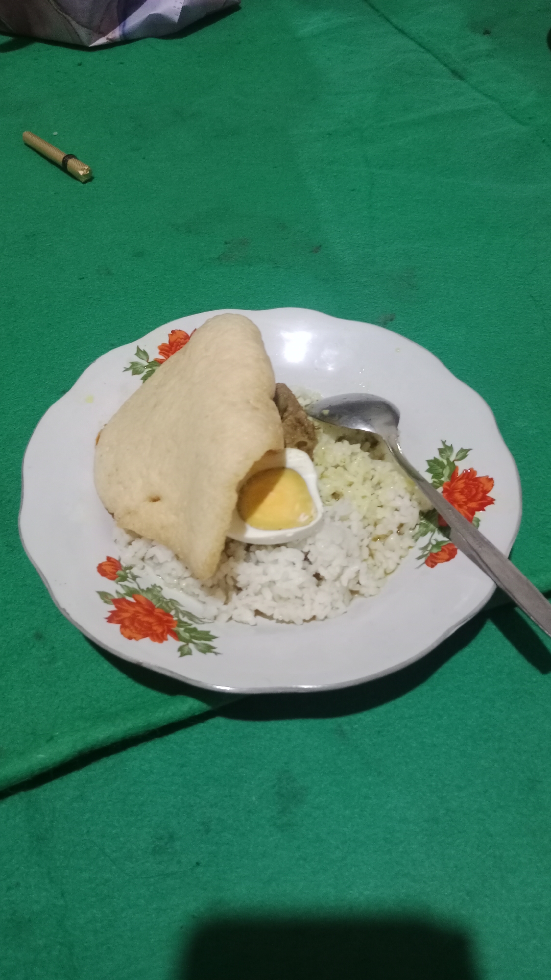 A plate of food sits on a green tablecloth. The plate is white with red and green floral patterns around the rim. On the plate, there is white rice topped with a piece of fried cracker, a hard-boiled egg cut in half, and a small portion of meat. A silver spoon rests on the right side of the plate. In the background, a small golden object, possibly a pen or pencil, lies on the green tablecloth. The scene appears to be indoors, with a shadow cast on the lower right of the image, suggesting artificial lighting. This image was taken in Pondok Kelor, Indonesia.