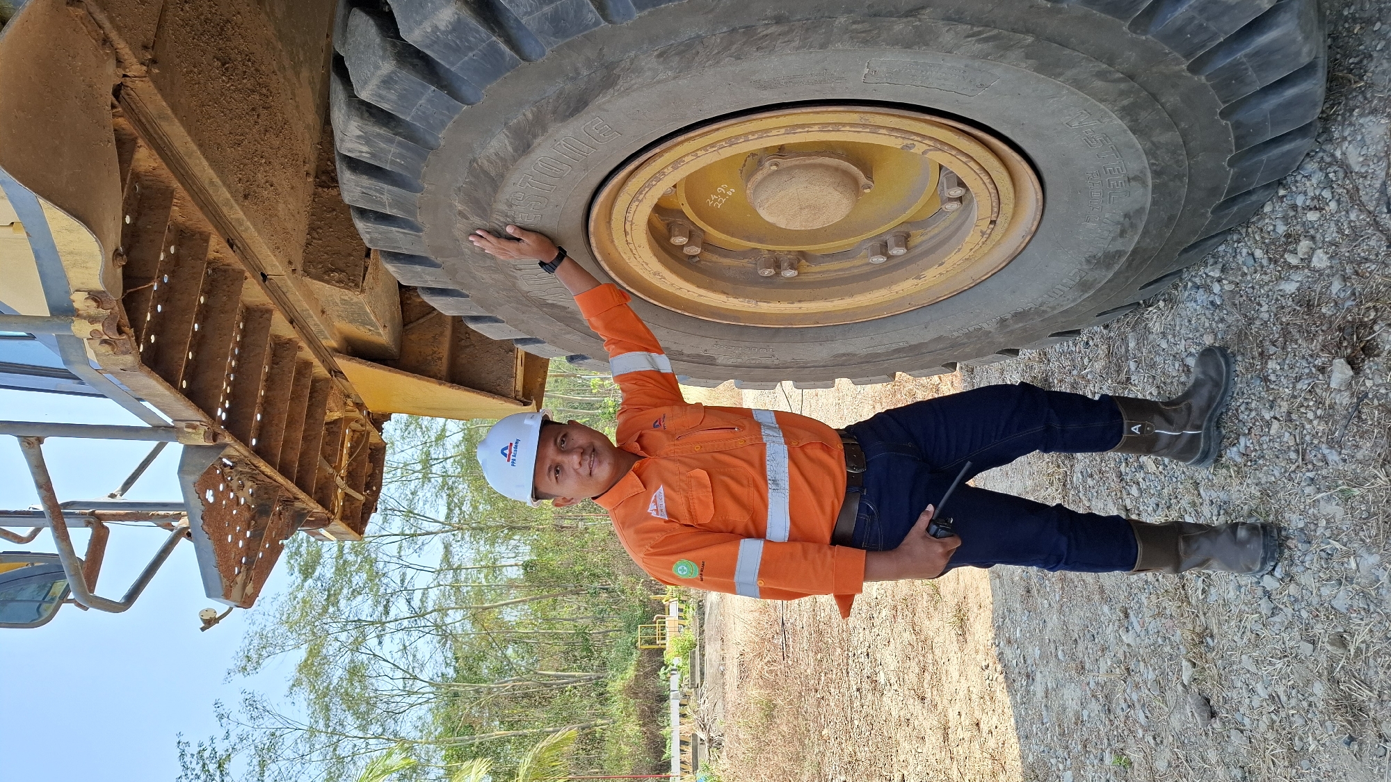 A man wearing a white hard hat with an "FPA Academy" logo and an orange high-visibility jacket with reflective stripes stands next to the massive tire of a yellow mining truck. The man is holding a black radio in his left hand and has his right hand resting on the tire. He is wearing dark blue jeans and dark brown work boots. The ground is covered with gravel and dry vegetation. In the background, there are trees and a clear blue sky. The location is identified as Unknown, Indonesia.