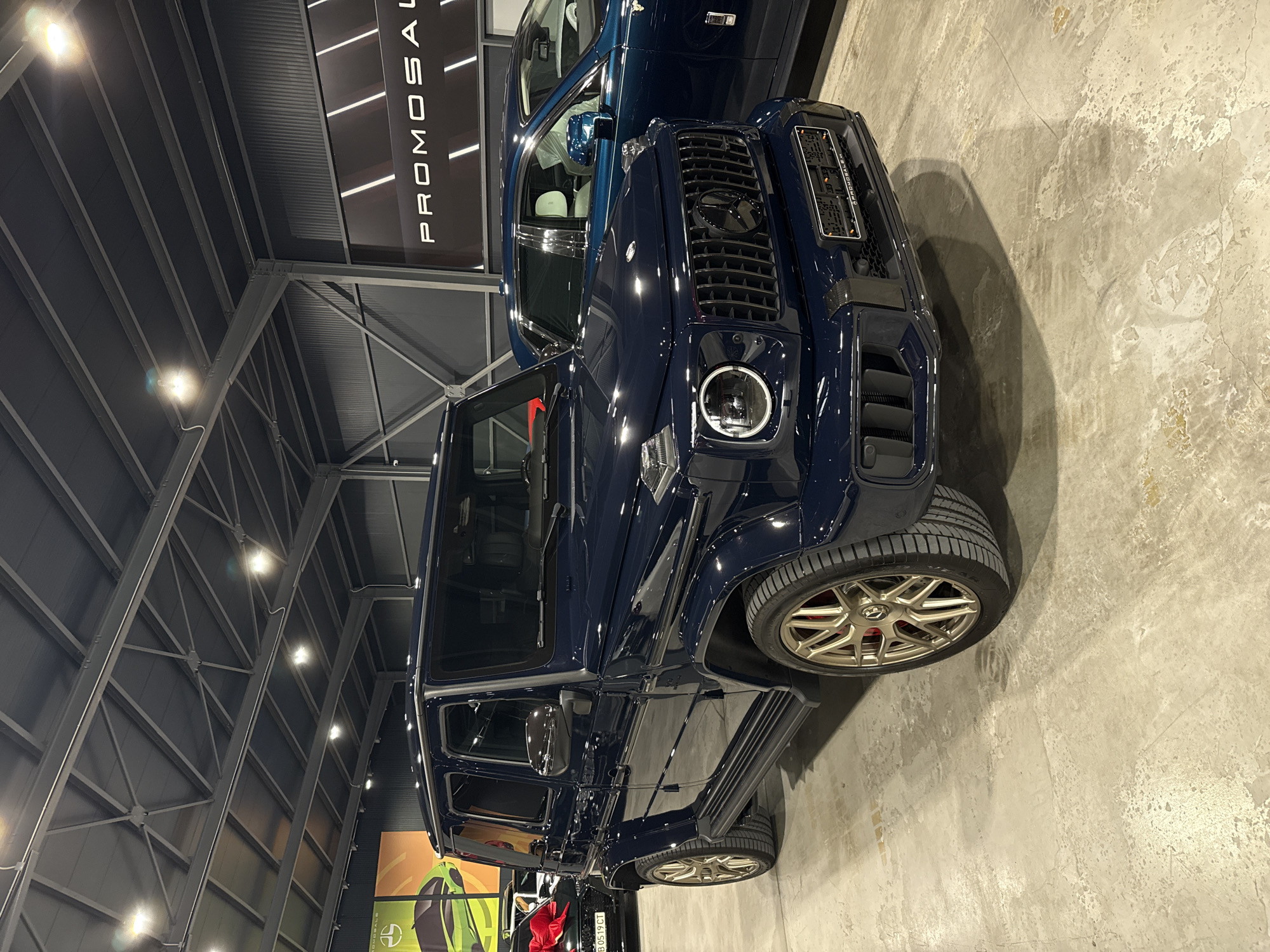 The image shows a dark blue Mercedes-Benz G-Class SUV in an indoor showroom with a concrete floor. The car is parked next to another vehicle, partially visible behind it. A sign reading "PROMOSALE" is visible in the background, mounted on the showroom's metal frame structure. The showroom appears to be located in an industrial building. While the exact location within Plovdiv, Bulgaria is not discernible from the image, the presence of the "PROMOSALE" sign suggests it could be an automobile dealership or a car customization shop. No people are visible in the image.