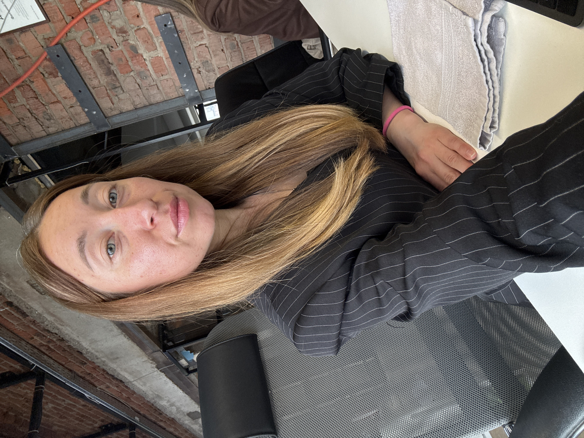 The image shows a woman with blonde hair, wearing a black pinstripe blazer and a pink wristband, seated at a white desk. A folded grey towel rests next to her hands on the desk. She is in a room with an exposed brick wall and ceiling, featuring metal supports. The chair is a mesh-backed office chair. The location in the city of Chaikovsky, Russia, cannot be verified from the image.