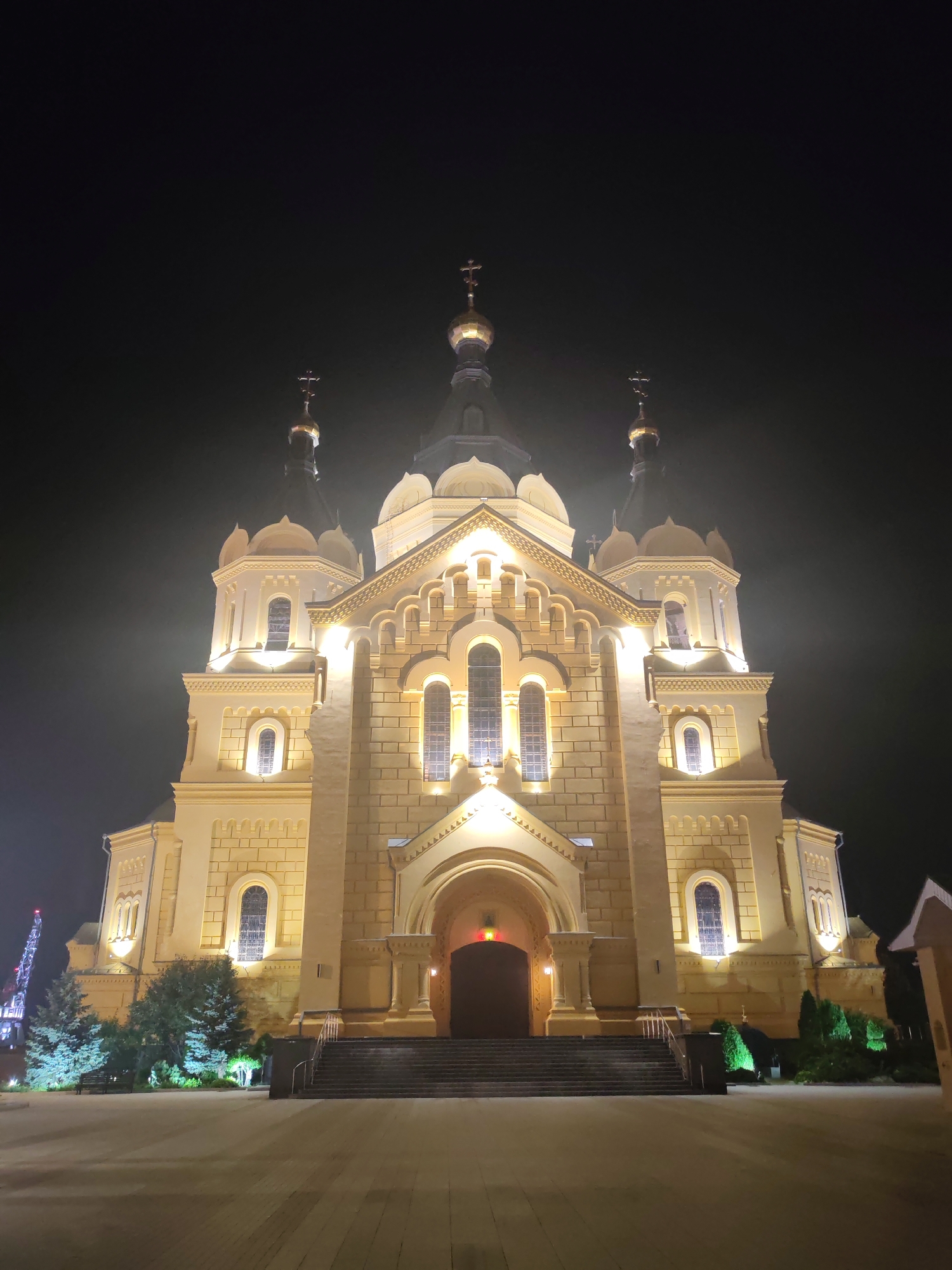 The image depicts a large, illuminated church at night, likely the Cathedral of Saints Peter and Paul, located in Nizhny Novgorod, Russia. The church is constructed of light-colored stone and features multiple domes topped with crosses. The facade is ornate, with arched windows and intricate architectural details. Warm yellow lights illuminate the building, creating a striking contrast against the dark, misty sky. A wide, paved courtyard is visible in the foreground, with steps leading up to the main entrance of the church. To the left of the church, several trees are present, and in the far distance, a large industrial structure with lights is visible. The overall atmosphere is one of solemnity and grandeur.