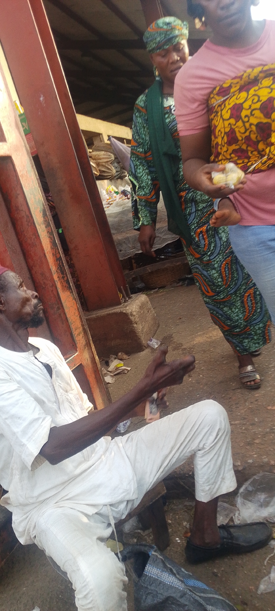 This image captures a vibrant market scene in Lokoja, Nigeria, featuring several individuals and market infrastructure. In the foreground, an older man with a beard, dressed in a white traditional top and trousers, sits on a low wooden stool. He is looking upwards and to the right, holding a small, light-colored object in his right hand. A black shoe and a blue and grey checkered bag are visible on the ground near his feet, amidst scattered debris like plastic wrappers.
Behind and to the right of the man, two women are partially visible. One woman wears a colorful green and orange patterned headwrap and a matching long-sleeved garment, possibly draped with a green scarf. Her face is slightly blurred but appears to be looking forward. Adjacent to her, another woman's torso and legs are visible; she wears a pink short-sleeved top, light blue jeans, and a yellow and red patterned fabric, possibly a baby carrier. She holds a small, wrapped item, likely food, in her hands. She wears light-colored sandals.
Dominating the left side of the frame are large, reddish-brown metallic structures, likely supports for market stalls or a building. The background reveals a busy market environment with indistinct market goods, baskets, and debris on the ground, under what appears to be a basic roof structure. The scene is brightly lit, suggesting daytime. No discernible text is visible in the image.