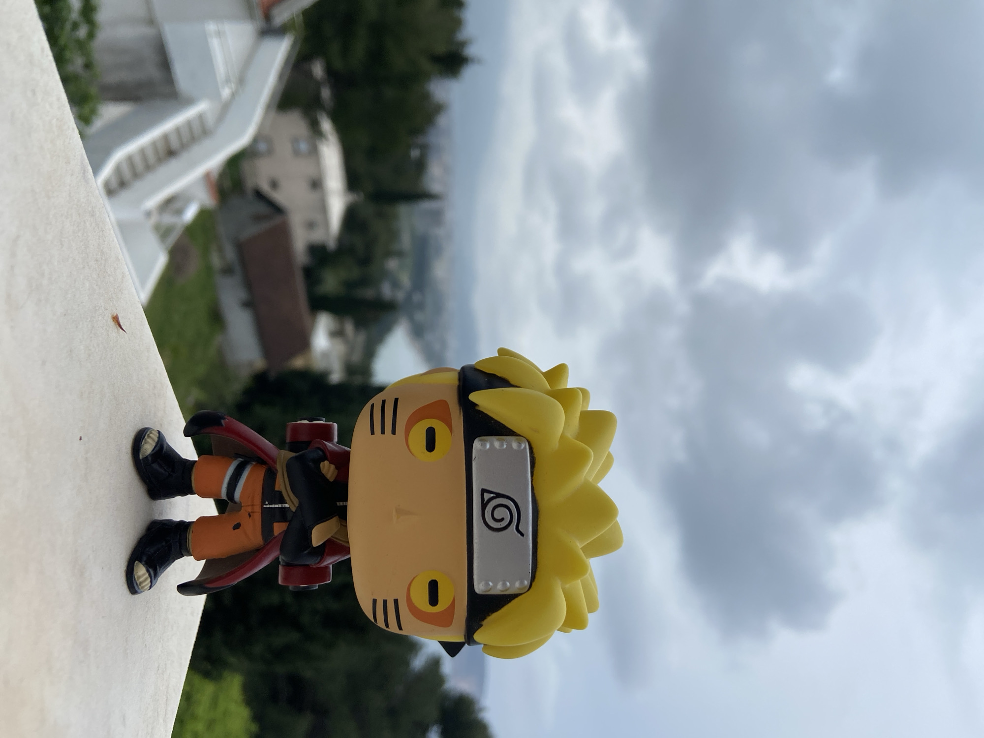 The image, rotated 90 degrees clockwise, presents a Funko Pop figure of Naruto Uzumaki standing on a light-colored, textured ledge or balcony. The figure is centered and in sharp focus, while the background is softly blurred.
Naruto is depicted in his iconic orange jumpsuit with black accents, black ninja sandals, and a black jacket or cape with a red lining. His distinctive spiky yellow hair is prominent, and he wears a black headband with a silver plate on his forehead, which features the embossed Konoha (Leaf Village) symbol—a spiral with a line through it. His face has stylized orange eyes with black pupils and three black whisker-like markings on each cheek.
The blurred background reveals a distant landscape under an overcast, cloudy sky. Faint details suggest a town or settlement with various buildings, some appearing white with visible staircases, nestled among green trees. A hazy, grey-blue body of water, likely the sea, is visible in the mid-ground, indicating a coastal environment. The sky is filled with soft, grey clouds, suggesting an overcast daytime. The scene overlooks the coastal town of Šušanj, Montenegro.