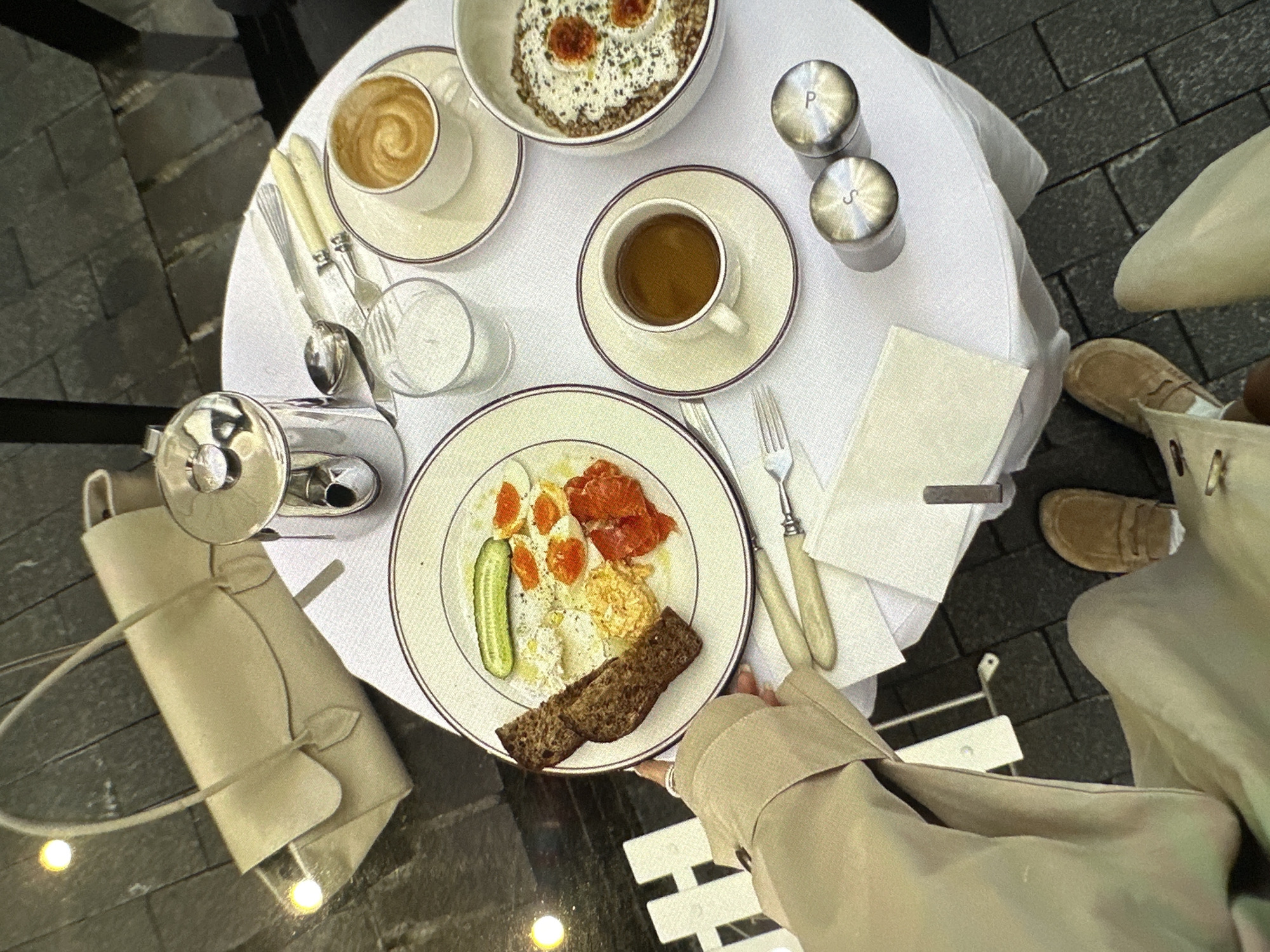 A high-angle shot reveals a round table set with breakfast items, situated on a paved surface in Yerevan, Armenia. A person wearing a light brown trench coat is seated at the table. The table is covered with a white cloth and holds a plate of food containing bread, cucumber, tomatoes, eggs, and other unidentified items. A bowl of porridge-like food and two cups of coffee are also present. Cutlery, salt and pepper shakers, a napkin, and a small pitcher complete the table setting. A light-colored handbag is hanging off the side of the table. The person's tan-colored shoes are visible near the edge of the table. The background shows a paved walkway with varying light conditions.