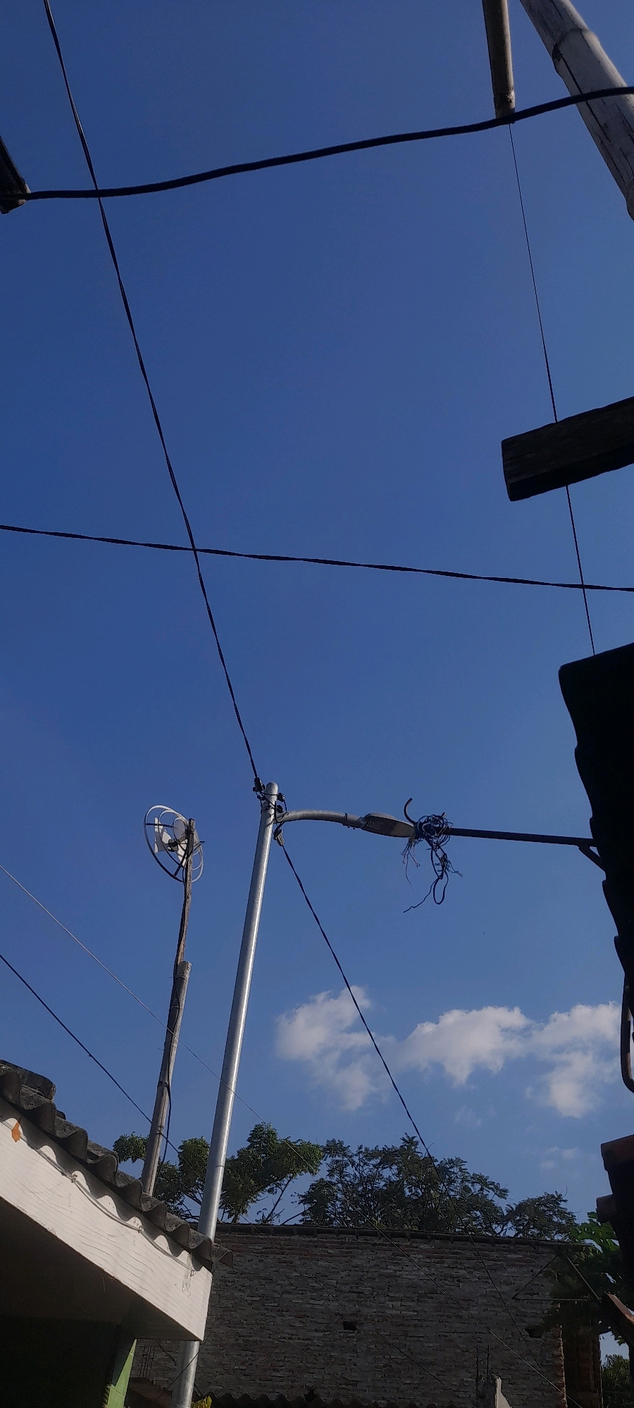 The image depicts a clear blue sky above a residential area in Tegal, Indonesia. Prominently featured are electrical wires crisscrossing the sky, a street lamp with tangled wires hanging from it, and a circular antenna mounted on a wooden pole. In the foreground, the corrugated roof of a white building is visible, as is a brick wall and trees. The scene appears to be an outdoor environment during the daytime.
