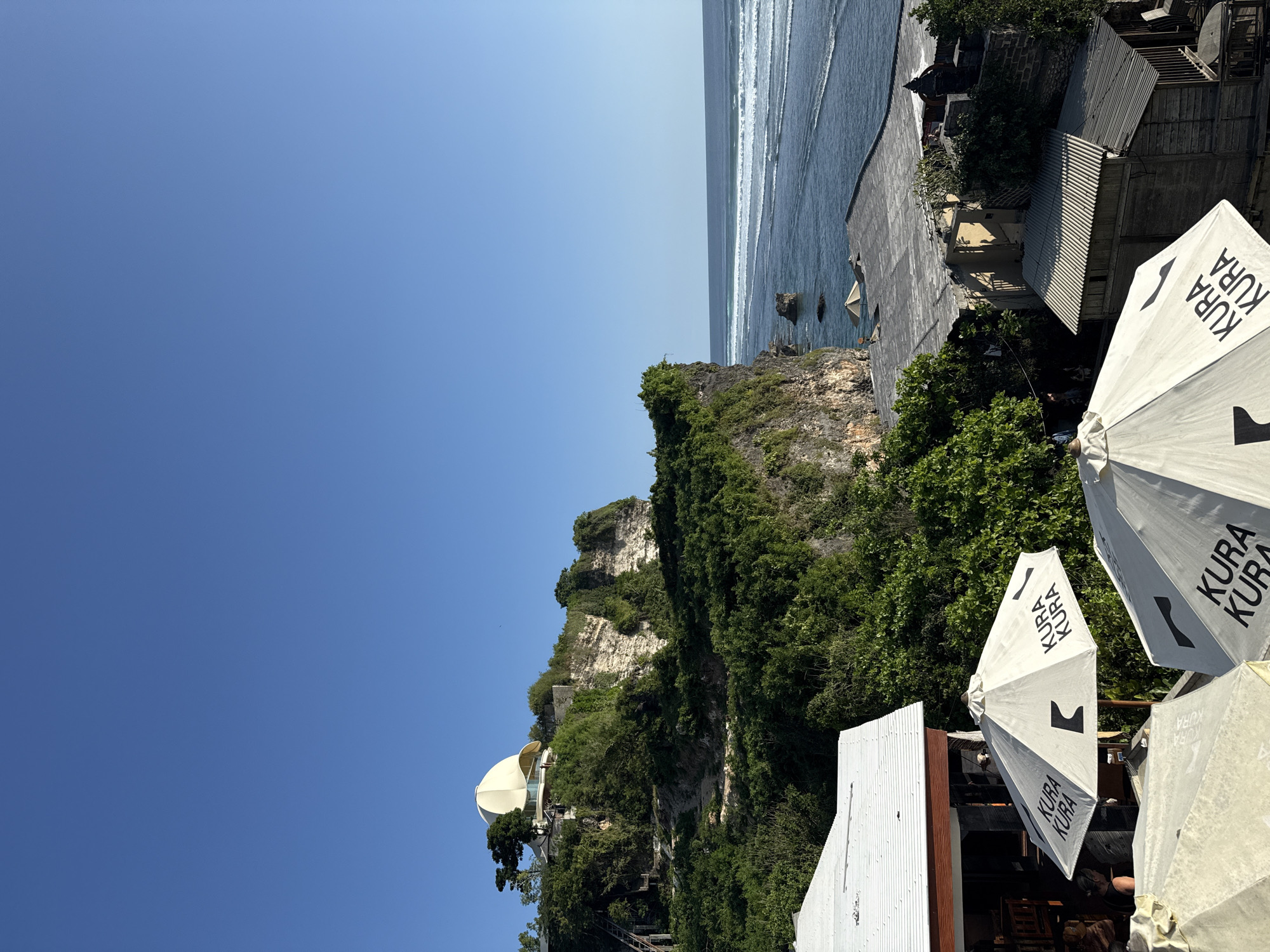 The image depicts a coastal scene, potentially at a cliffside location in Kuta Selatan, Indonesia. Clear blue sky dominates the upper left. The sea is visible at the top right, with waves approaching the rocky shore or a built-up area along the cliff edge. A rocky cliff face covered in dense green vegetation occupies the central portion of the image. The lower right shows features indicative of a commercial or tourist area, including white umbrellas with the word "KURA" on them, along with structures with metal roofing. The specific activities or interactions of people are not readily apparent from the image, but there are signs of human presence and development in the setting. A prominent white, dome-shaped structure sits on the cliffside surrounded by vegetation, potentially a building or tourist attraction.