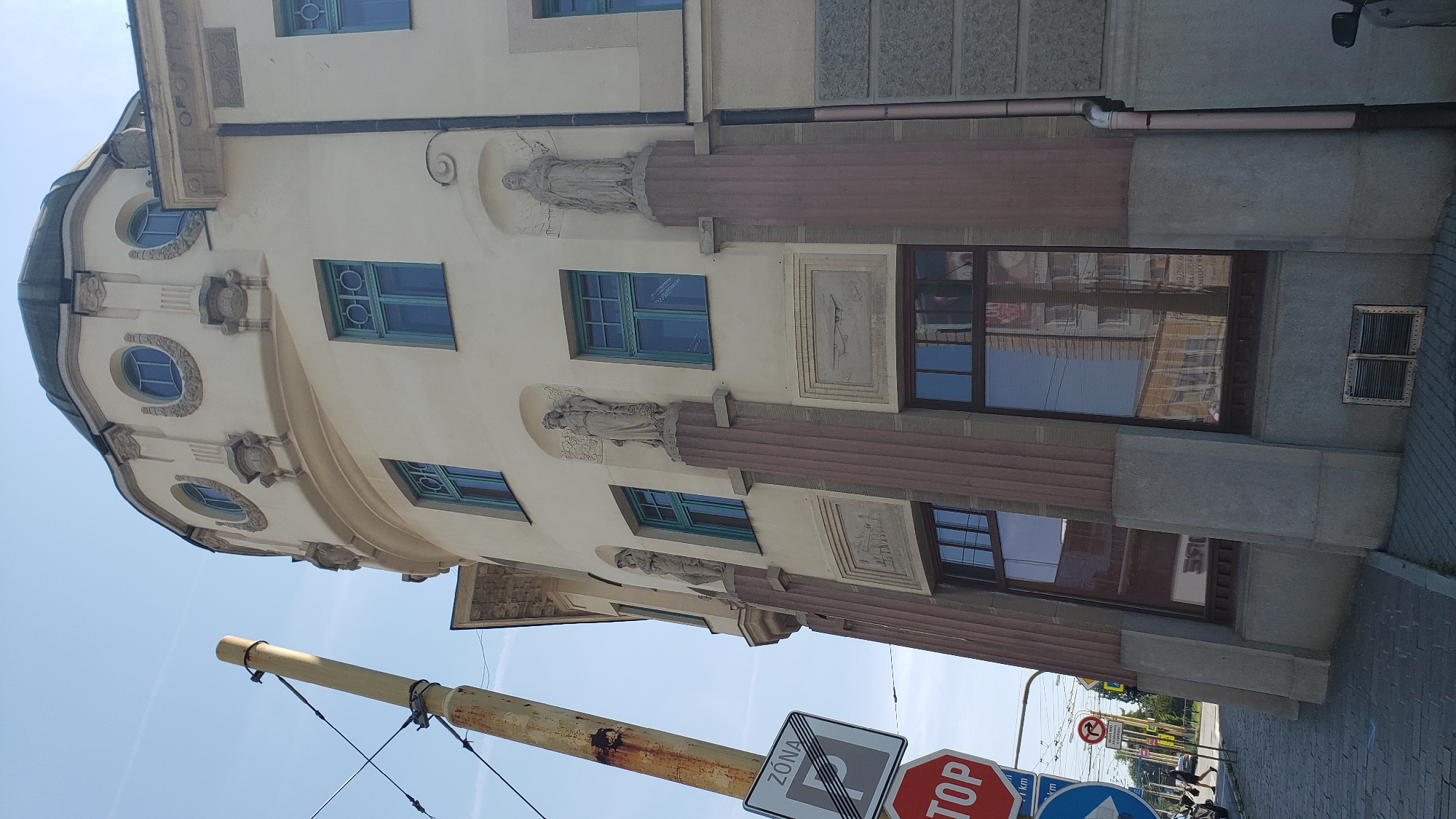 The image captures an exterior view of a historical building with a distinct architectural style, located in the city of Prešov, Slovakia. The building features a curved facade, a domed roof, and decorative elements such as statues and relief carvings. Several windows with turquoise frames are visible, interspersed with columns and alcoves. The building's facade is painted a light cream color. In the foreground, a yellow utility pole with overhead wires is prominently positioned on the left side of the frame. Road signs are also visible, including a "STOP" sign and a parking zone sign. The street in front of the building is paved with cobblestones, and there are indications of pedestrian activity in the background. The sky is clear and blue, suggesting a sunny day. Reflections of other buildings are visible in the large ground-floor windows. The overall scene depicts a blend of historical architecture and urban infrastructure.